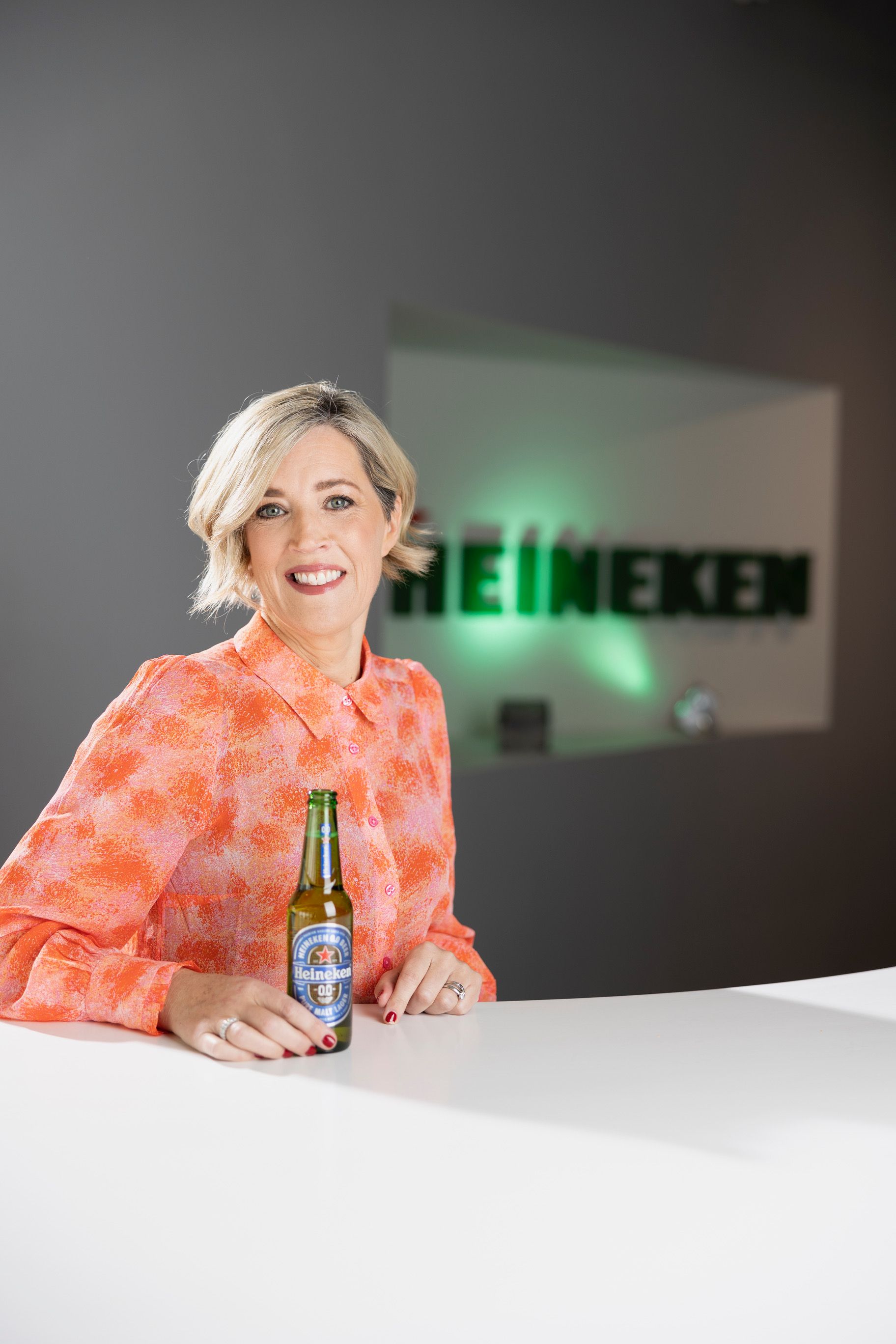 Woman in orange blouse holding a Heineken bottle, smiling, with the Heineken logo in the background.
