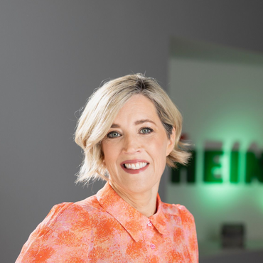 Woman with short blonde hair smiles, wearing an orange patterned shirt, in front of a Heineken sign.