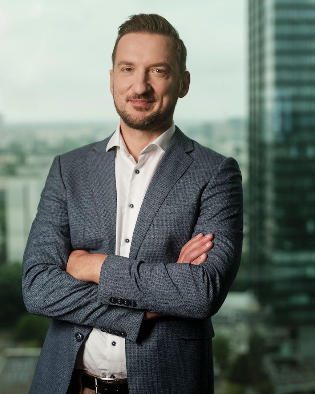 Man in a blue blazer with arms crossed, smiling. Set in an office with a cityscape in the background.