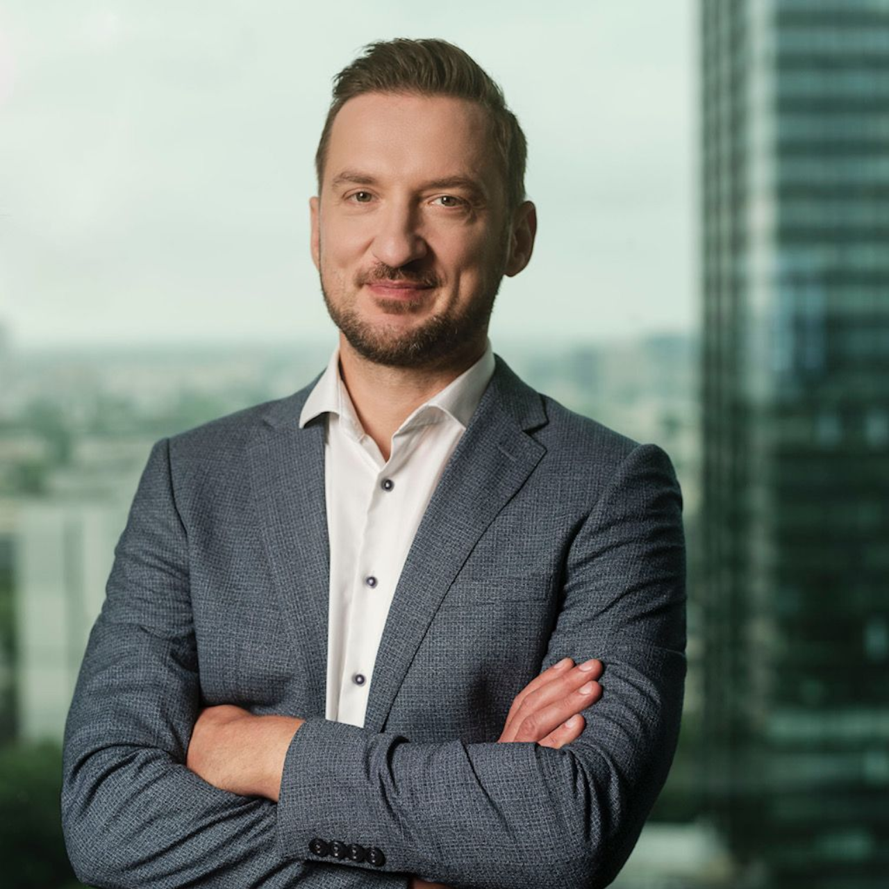 Man in a blue blazer with arms crossed, smiling. Set in an office with a city view.