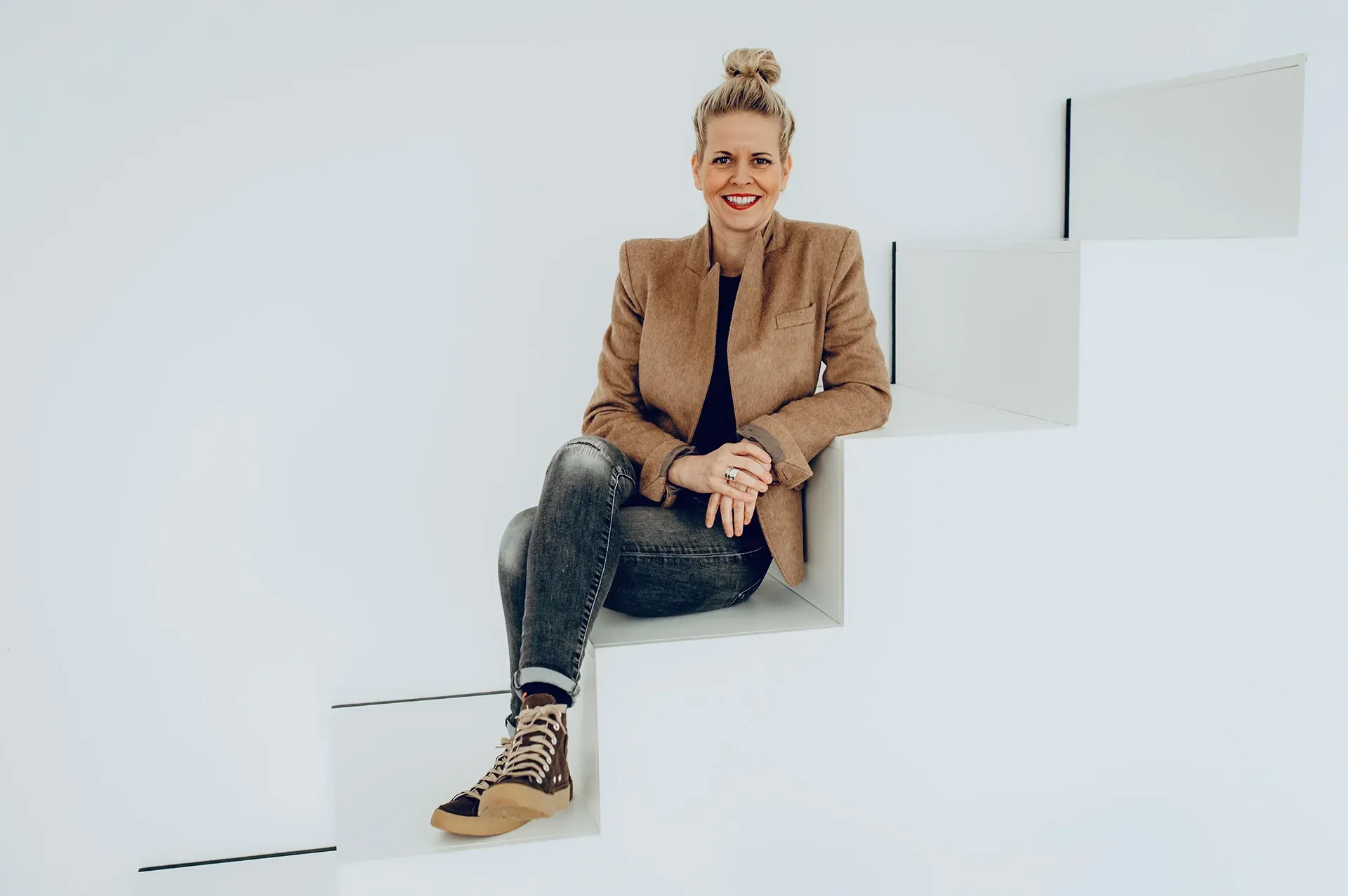 Woman seated on white block stairs, smiling. Wearing a brown blazer, dark jeans, and patterned boots.