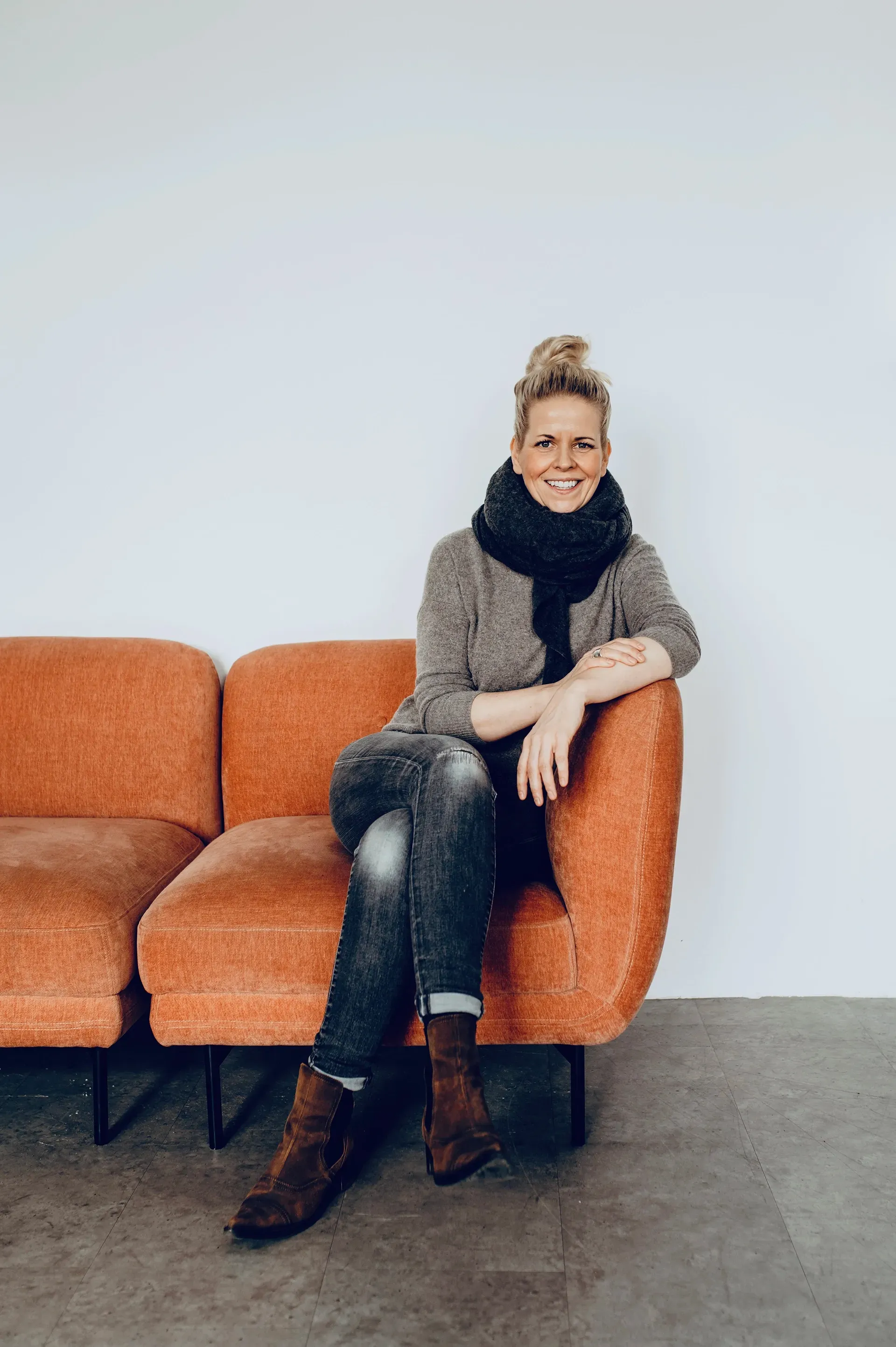 Woman on orange sofa, wearing scarf, blazer, and jeans, smiling.