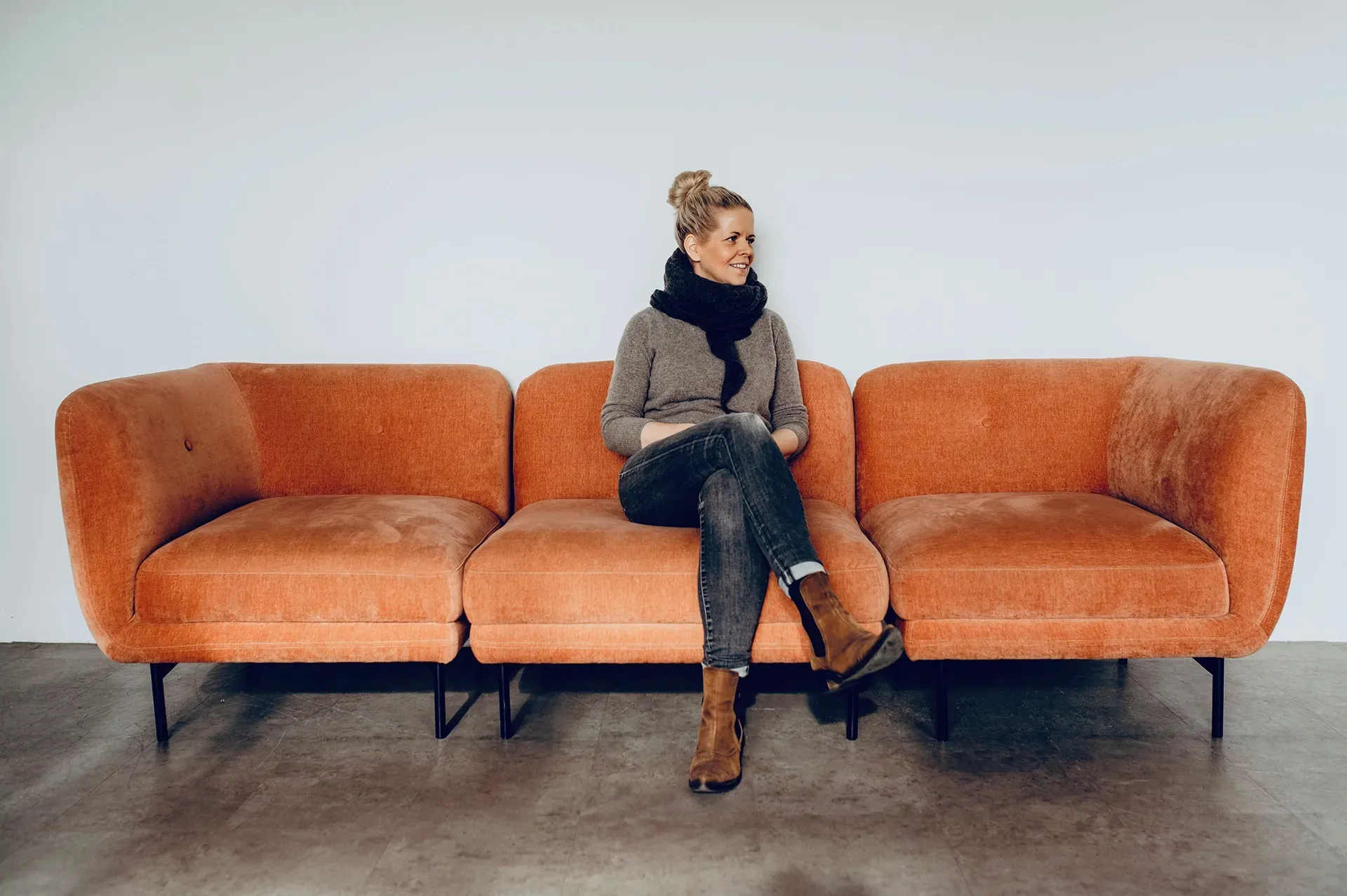 Woman sitting on an orange modular sofa, wearing a sweater, jeans, and a scarf, smiling.
