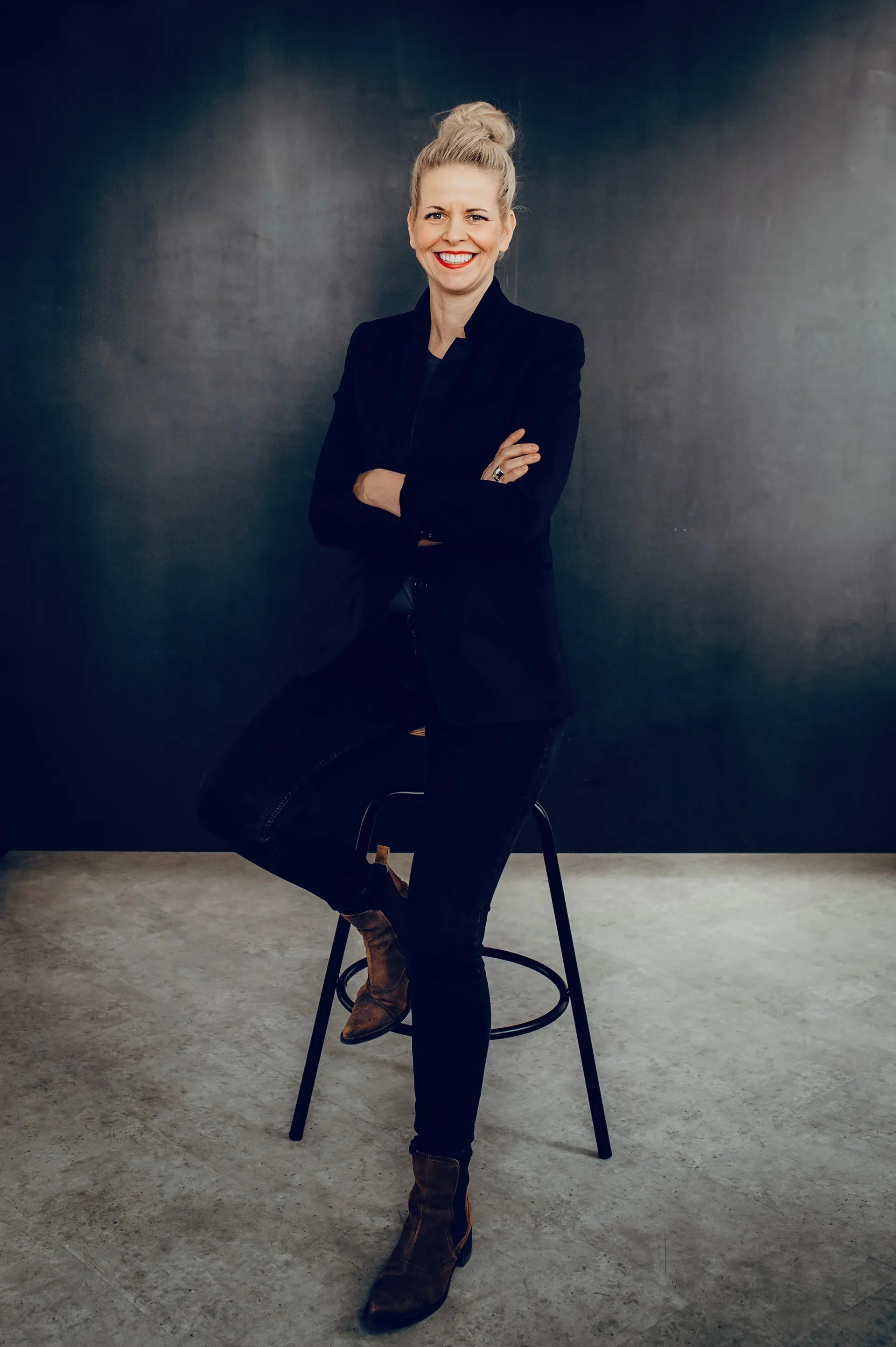 Woman with blonde bun smiles, arms crossed, seated on a stool, leaning against a dark wall.