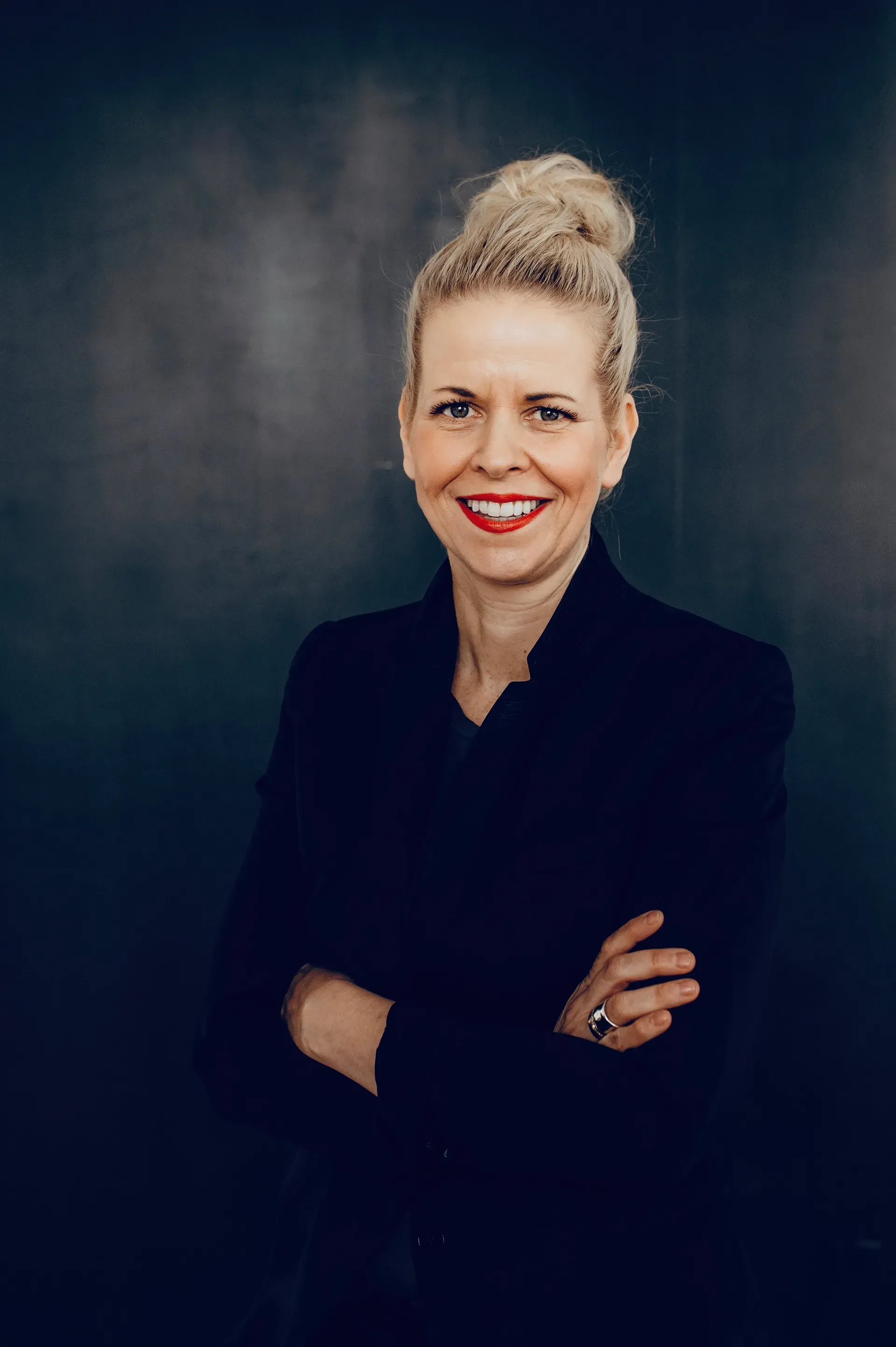 Woman in a black blazer with blonde hair in a bun smiles with arms crossed.
