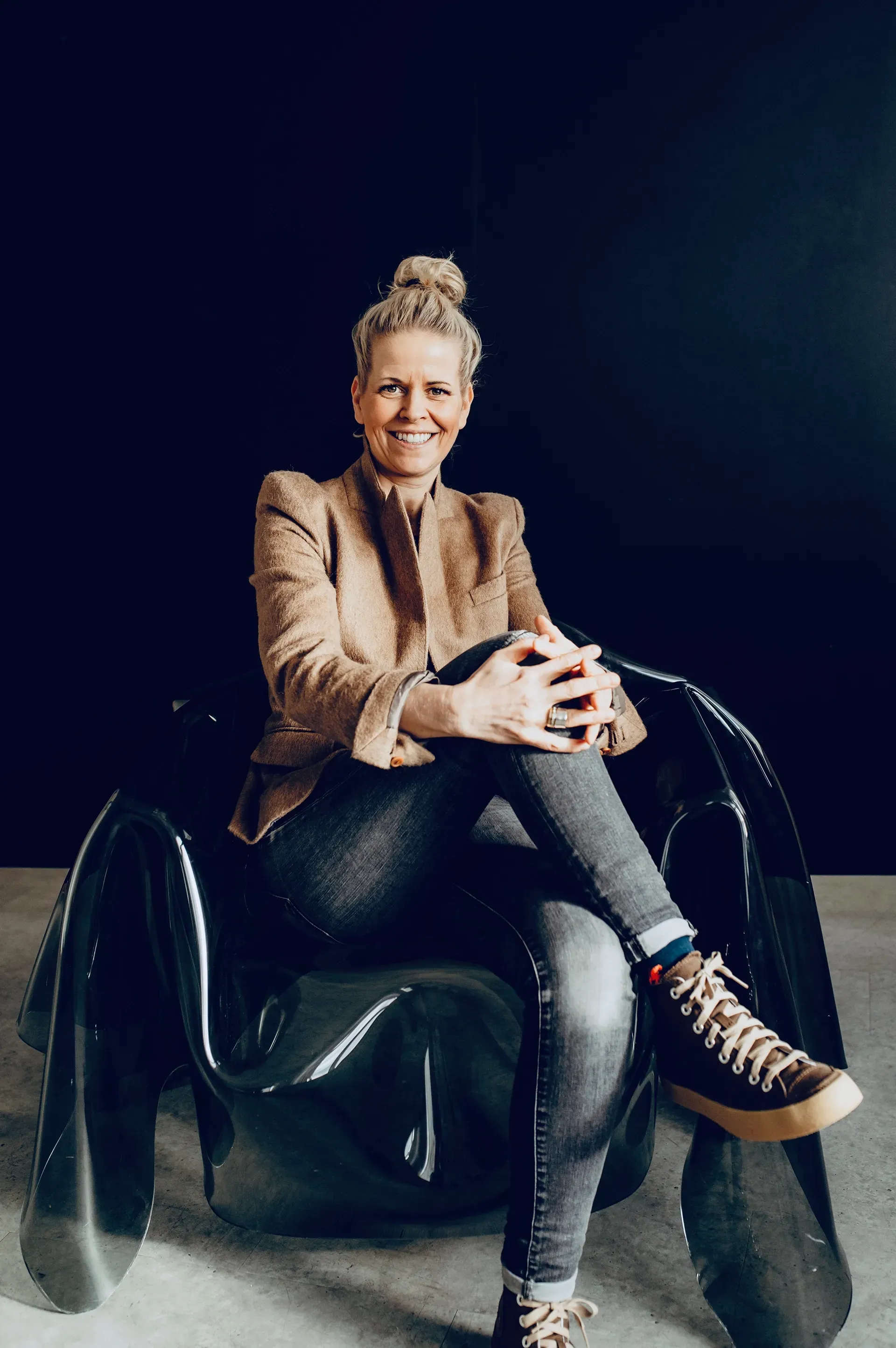 Woman in blazer and jeans sits smiling in a black, modern chair.