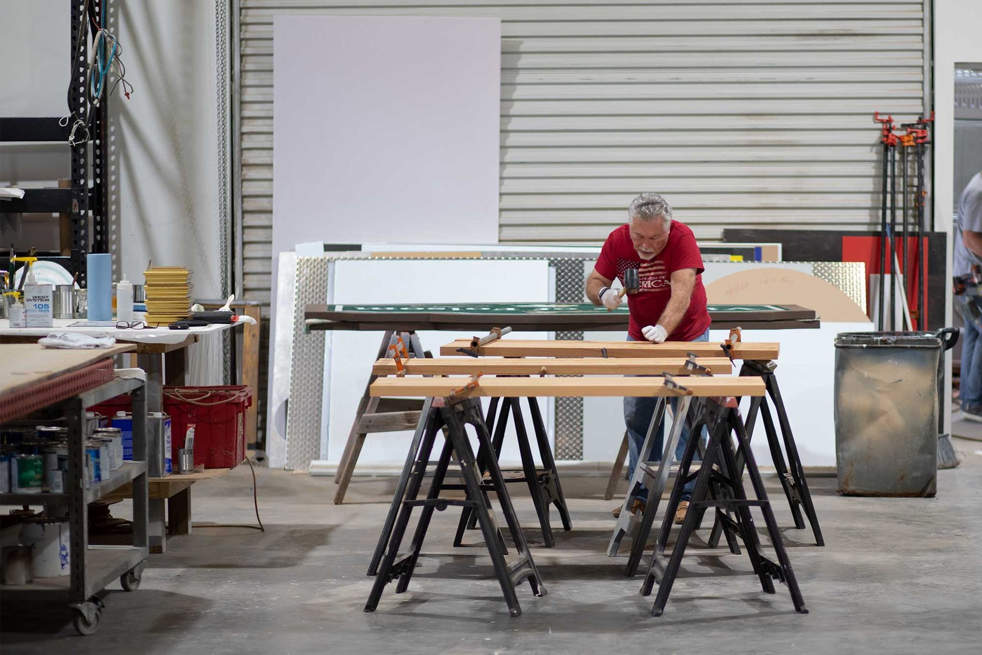 Man working on wood with clamps in a workshop. Sawhorses, tools, and paint are visible.
