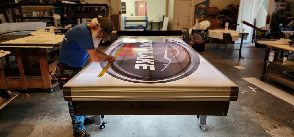 A man works on a large illuminated sign with a logo in a workshop.