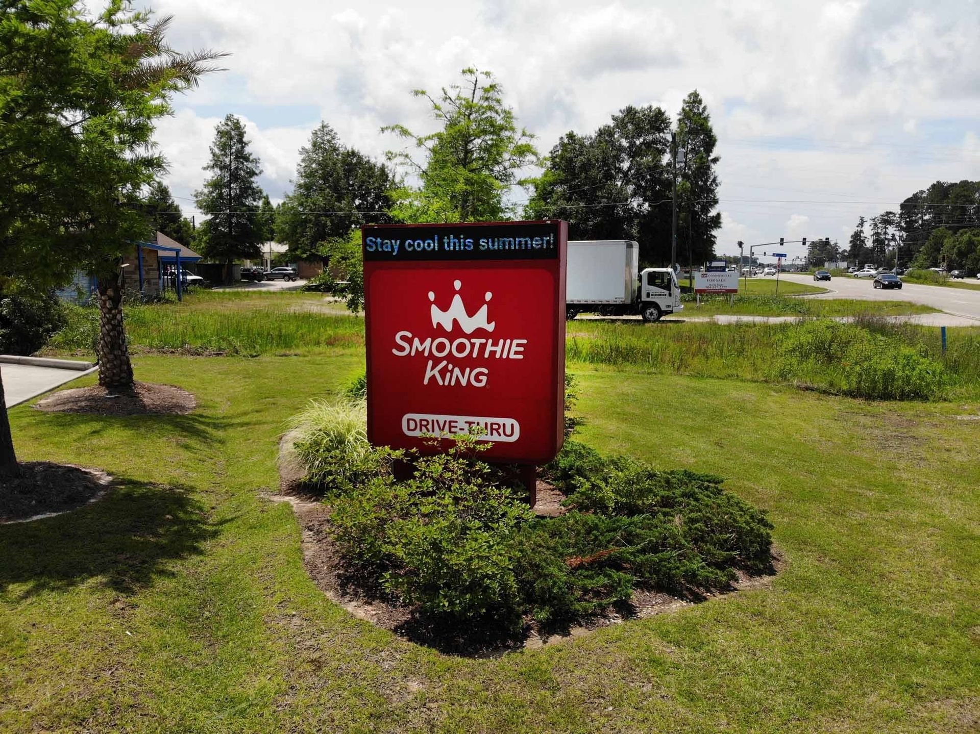 Smoothie King sign with red background, crown logo. Located in grassy area with a white truck in the background.
