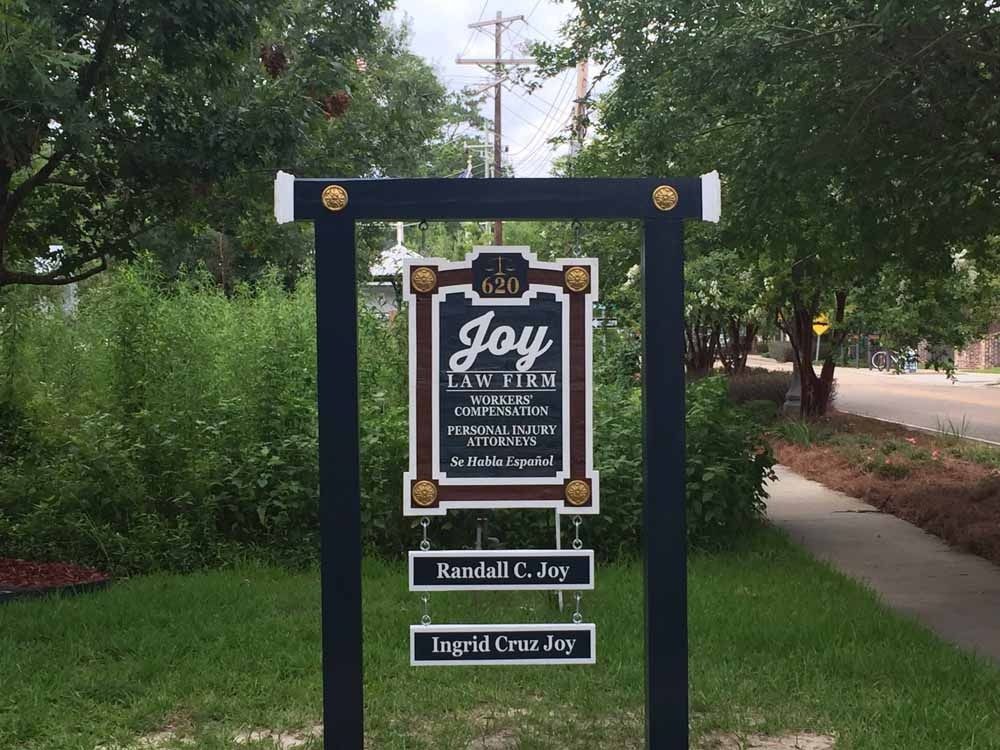 Sign for Joy Law Firm in front of a building with grass, trees, and a road. The sign has multiple layers and names.