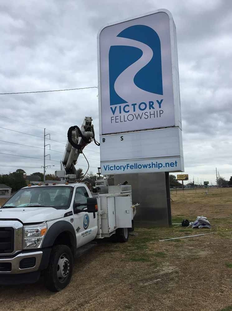 A work truck with a lift working on a tall sign for Victory Fellowship, under a cloudy sky.