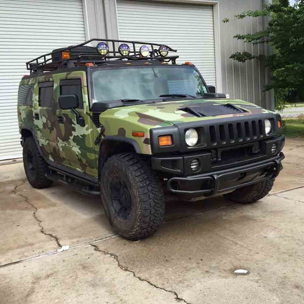 Camouflage-wrapped Hummer H2 SUV with black accents, roof rack, and off-road tires parked in front of a garage.