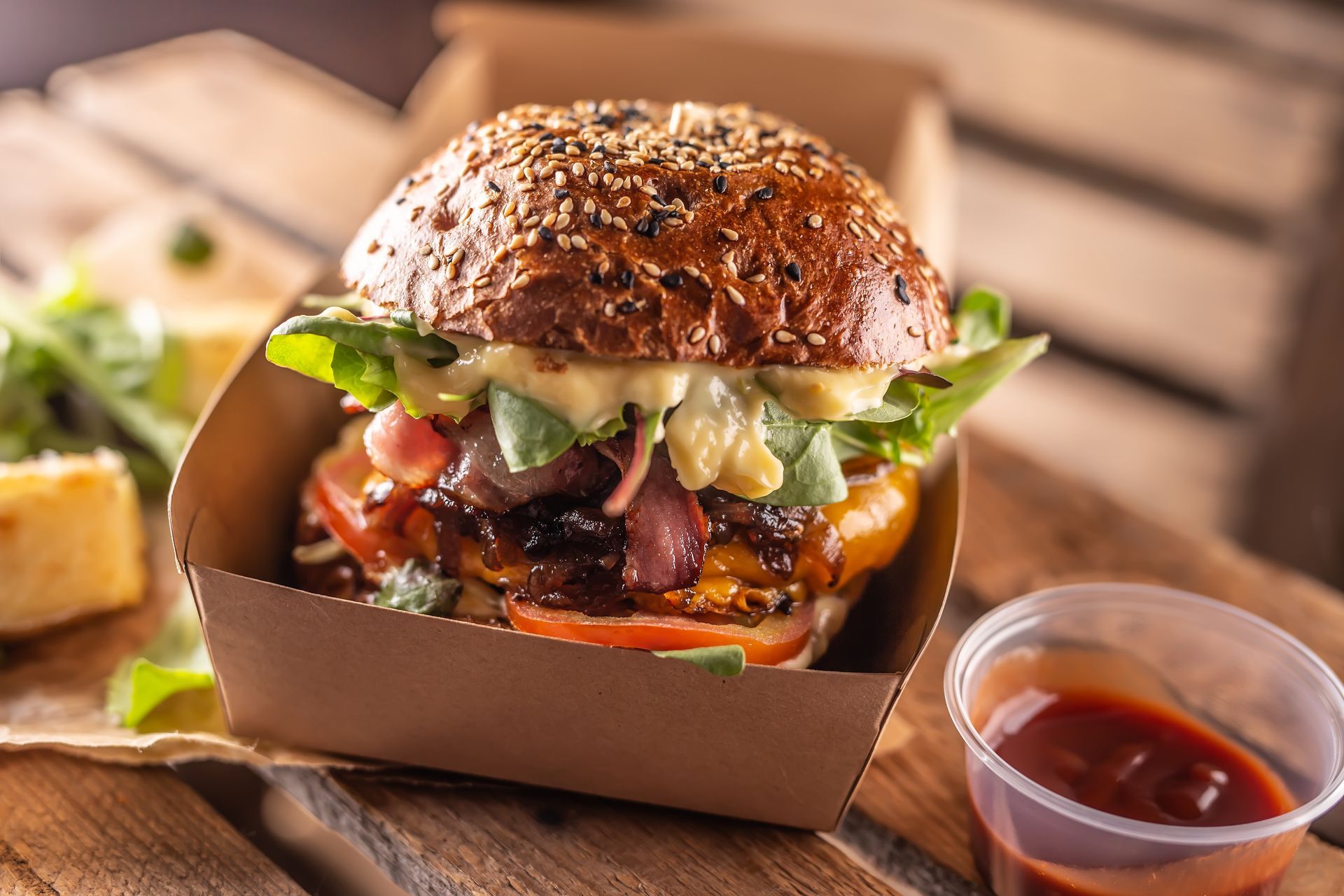 A hamburger is in a cardboard box on a wooden table next to a cup of ketchup.