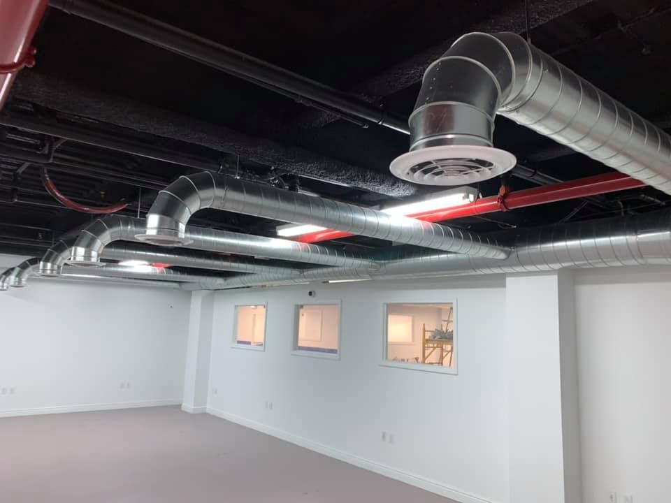Interior view of a room with exposed ductwork, pipes, and recessed lighting against a black ceiling. White walls and small windows are also present.