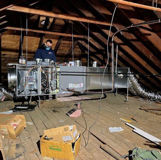 Person in an attic working on HVAC unit. Brown wood framing, cardboard boxes, and ductwork visible.