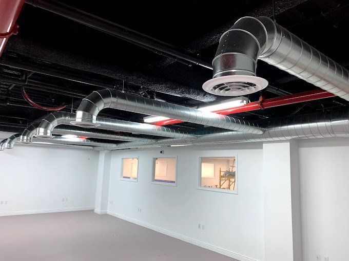 Silver HVAC ducts and lights on a black ceiling, in a white-walled room with small windows.