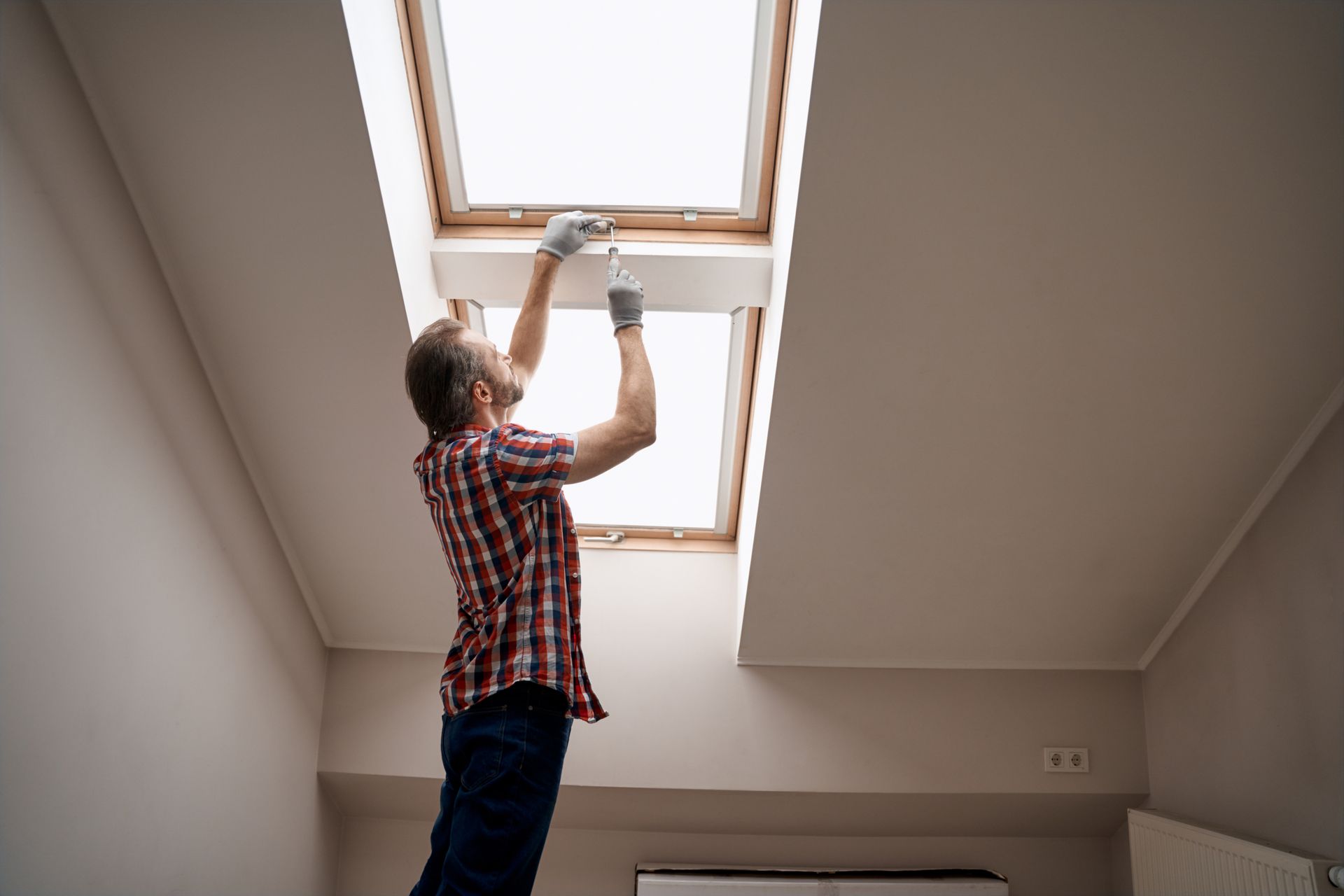 Man on a ladder installing something near two skylights. He wears gloves and a plaid shirt.