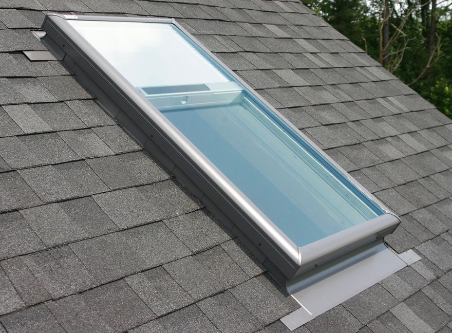 Skylight installed on a gray asphalt shingle roof. The window frame is metallic and holds a pane of glass.