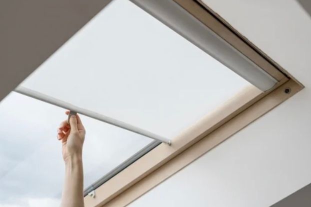 Hand pulling down a white skylight shade in a white ceiling with a wooden frame.