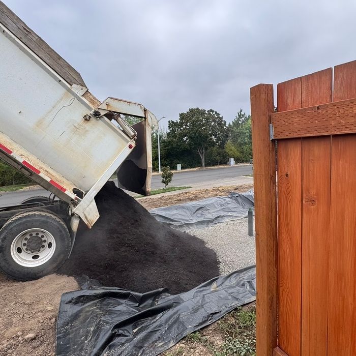 A dump truck pours a large pile of dark soil onto a black tarp in a yard next to a wooden fence.