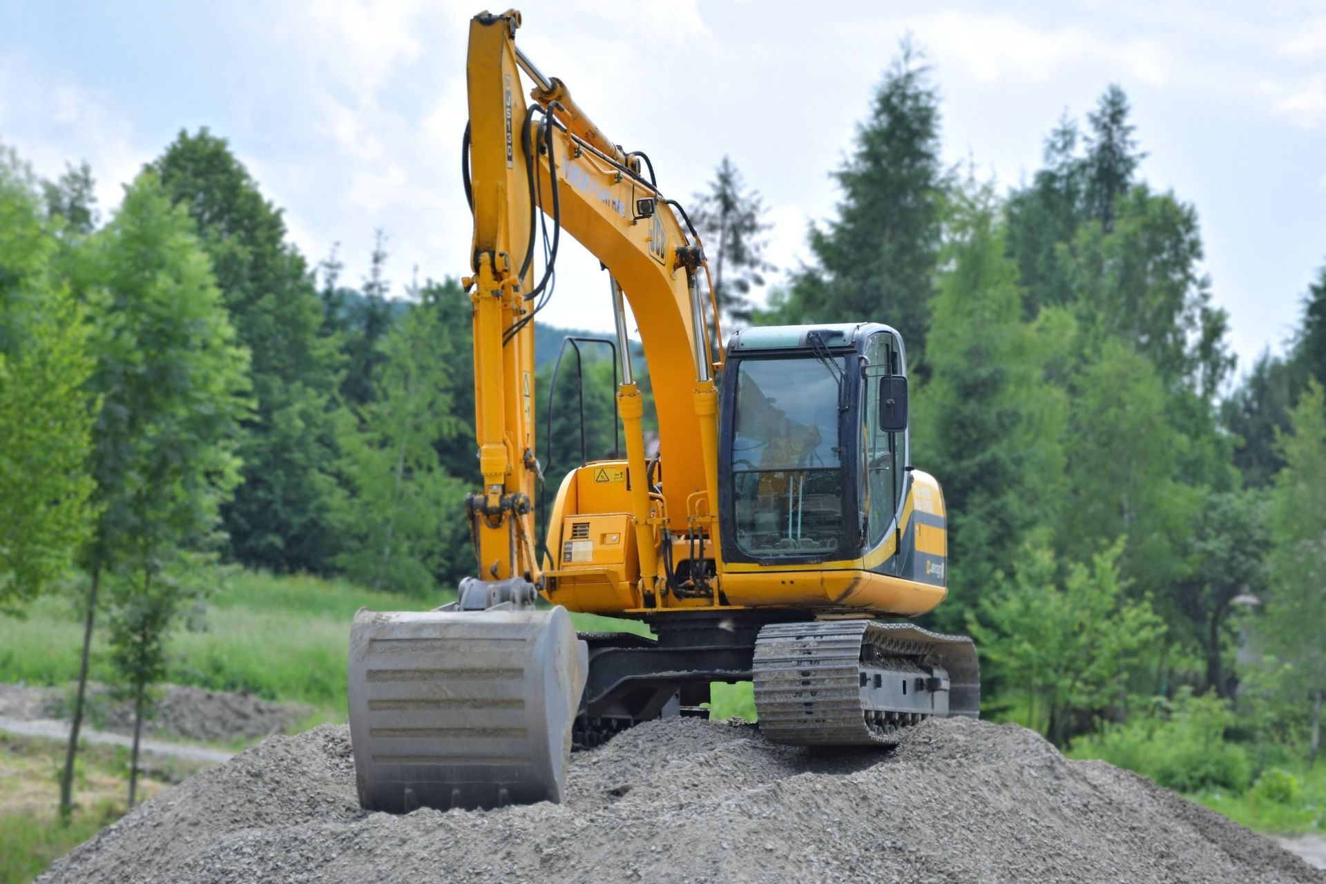 A yellow tracked excavator sits atop a large pile of gravel in a forested setting.