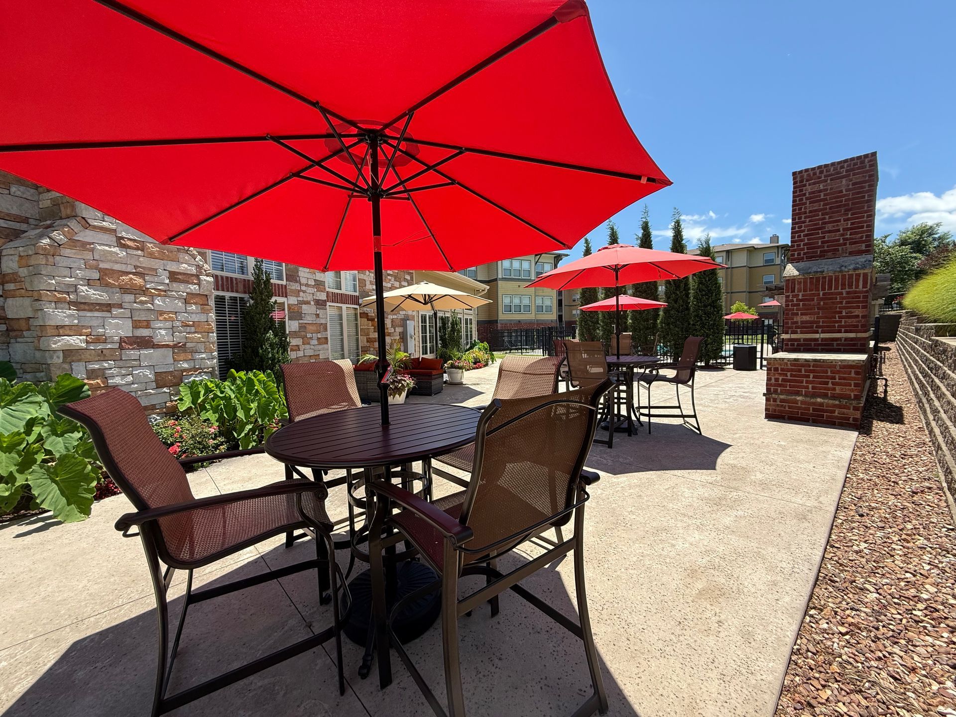 Outdoor patio area with brown metal tables and chairs under red umbrellas on a sunny day.