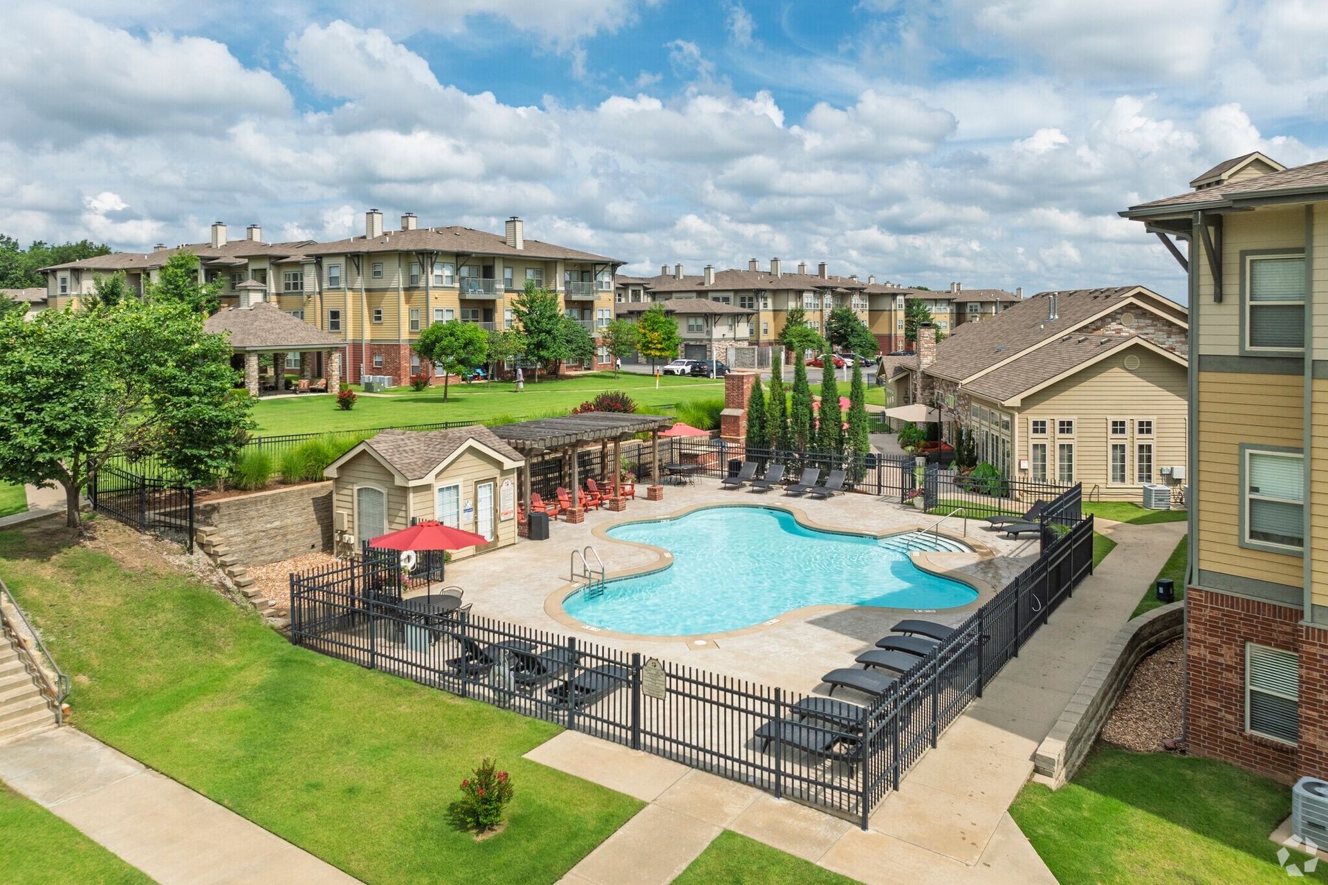 A sunny outdoor pool area at an apartment complex with lounge chairs, a pergola, and surrounding residential buildings.