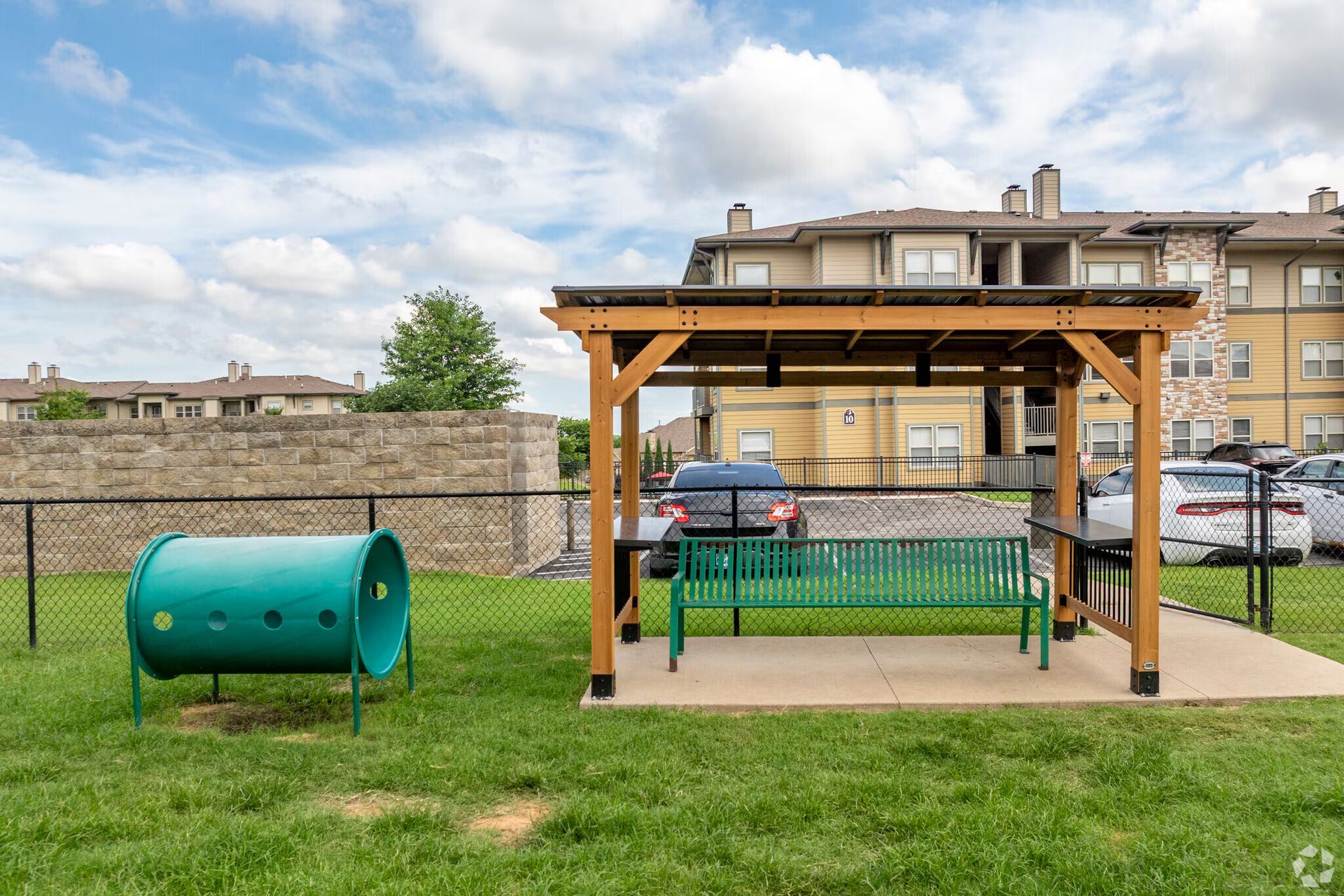 A green dog agility tunnel and a bench under a wooden pergola inside a fenced dog park at an apartment complex.