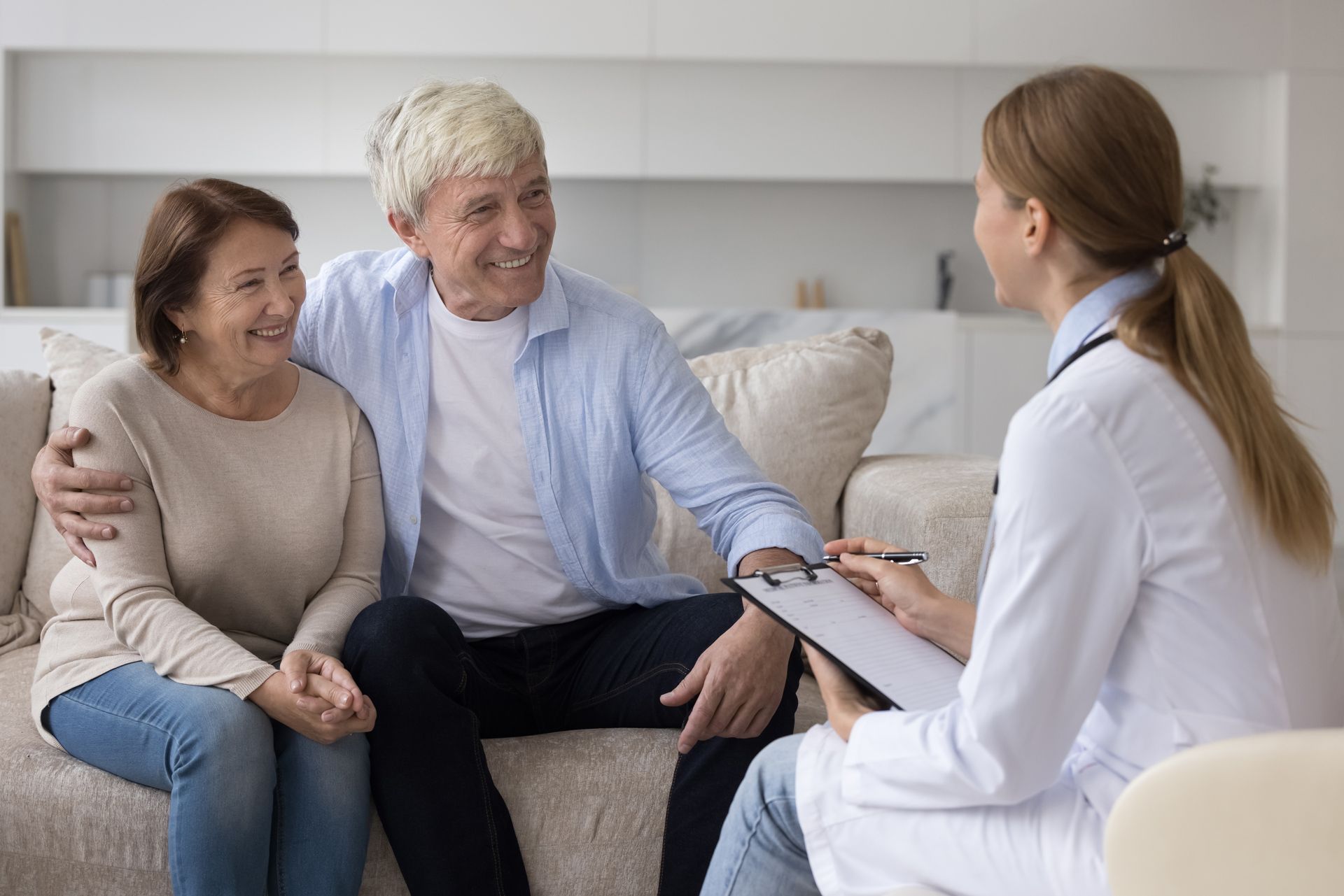 Couple on a couch consult with a doctor holding a clipboard in a well-lit room.