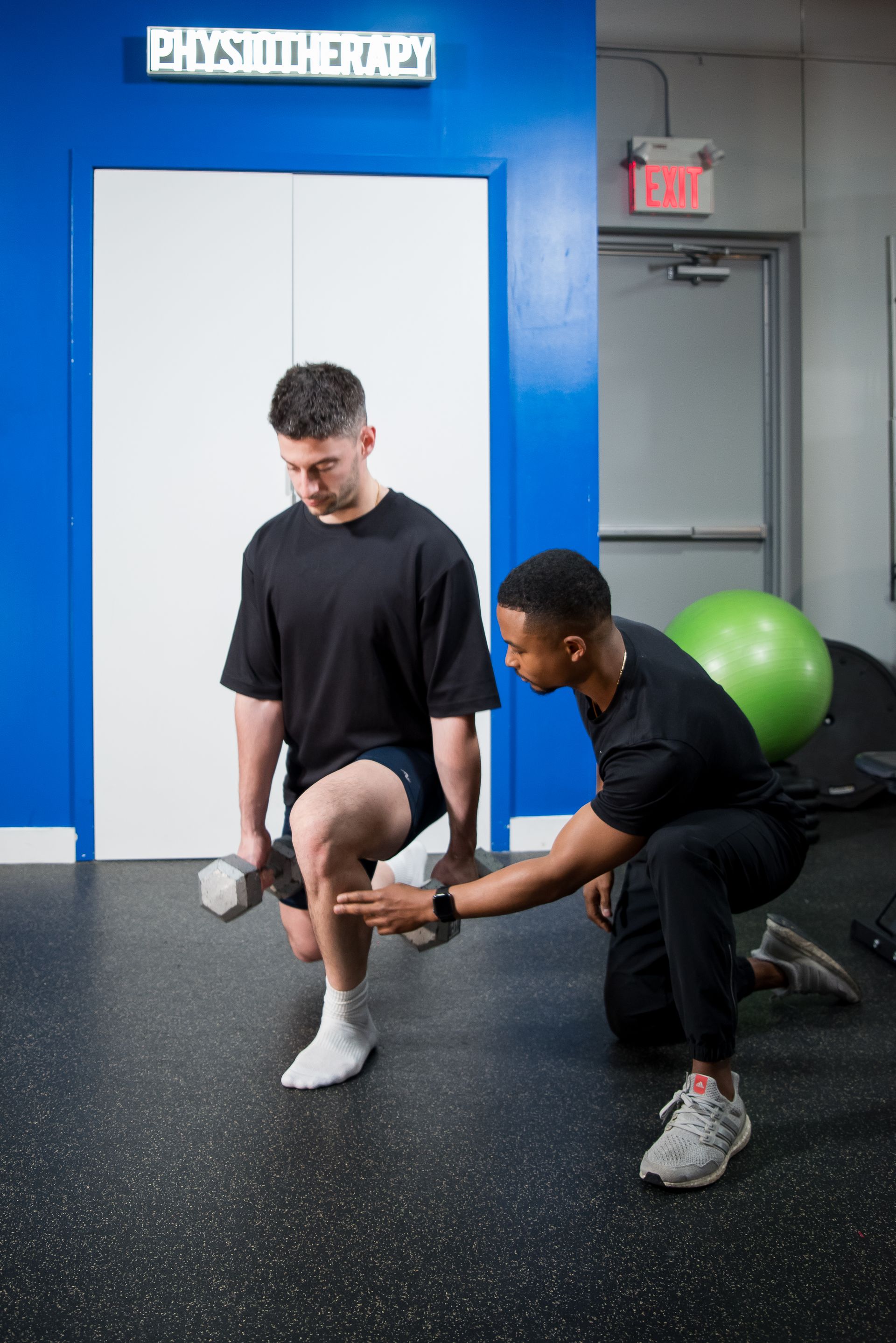 A physical therapist guides a person holding dumbbells through a weighted lunge in a clinic.