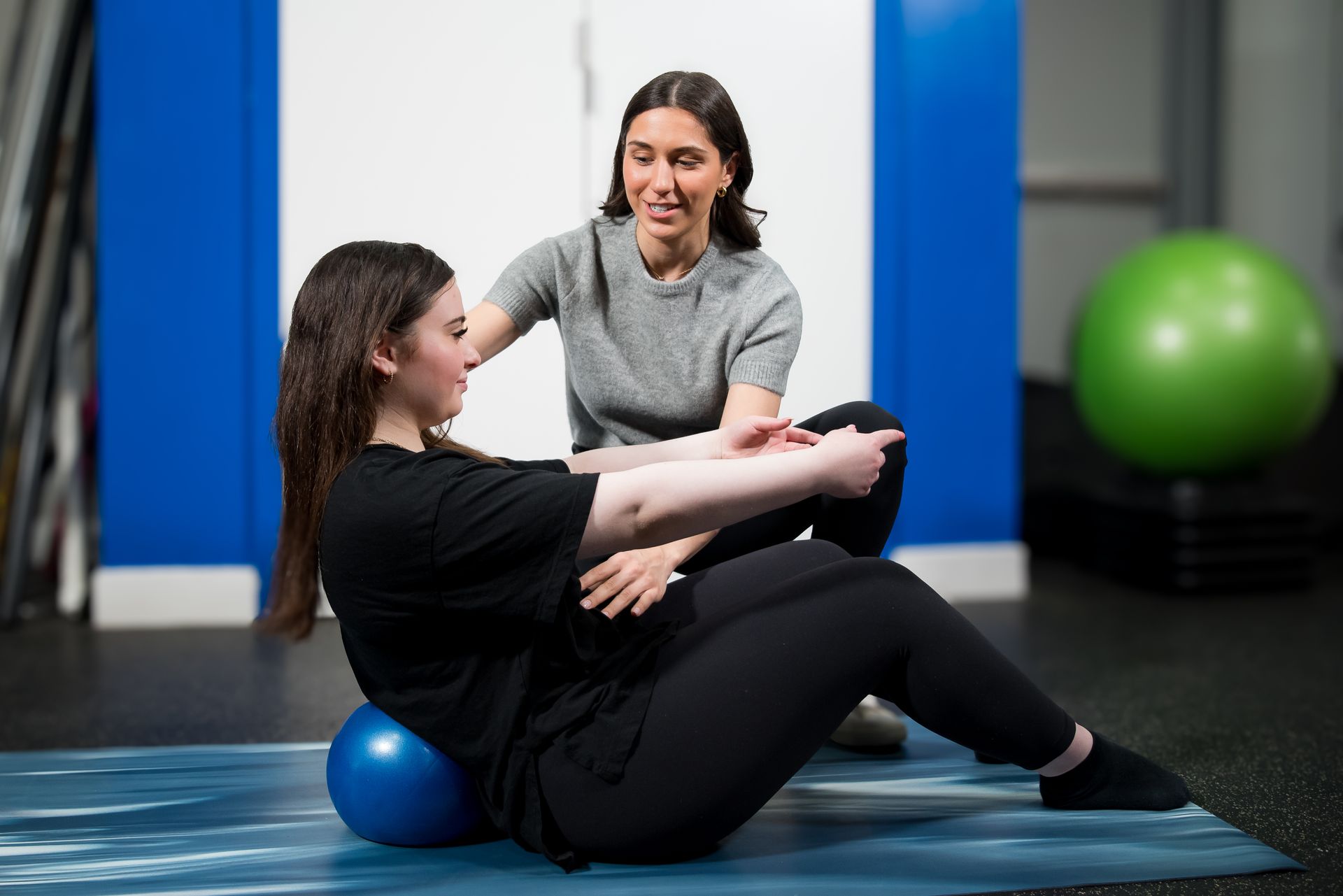 A physical therapist supports a person doing a core exercise on a floor mat using a small blue stability ball.