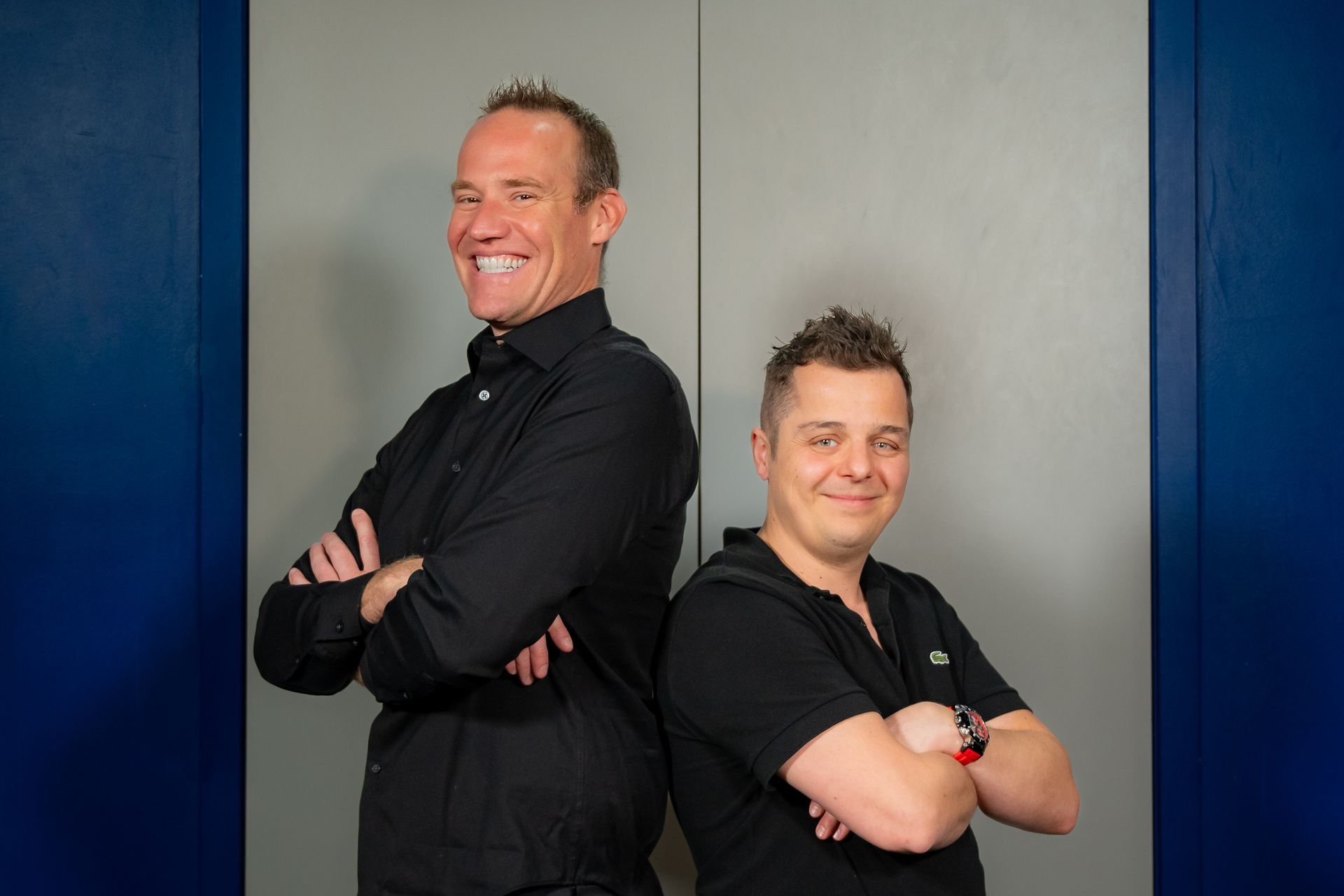 Two men stand back-to-back in front of a gray door with blue side panels, both smiling with their arms crossed.