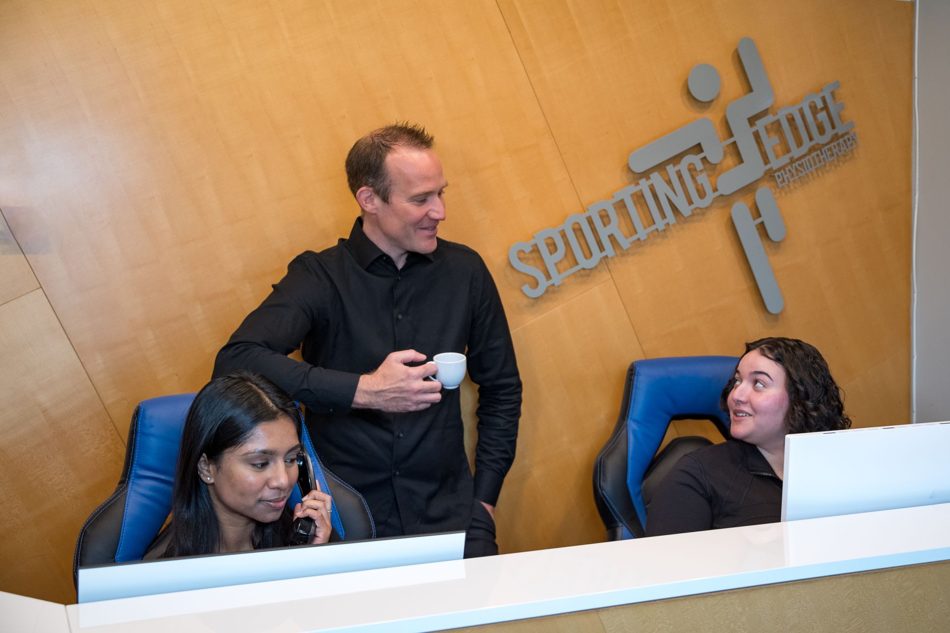 Two receptionists at a front desk, one using a phone and the other talking to a man standing beside them holding a cup.