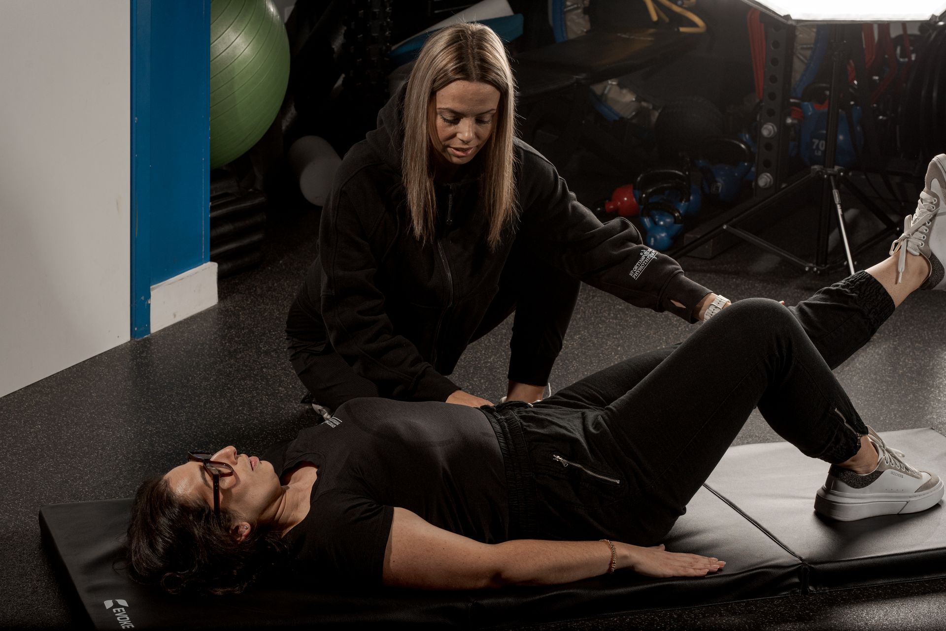 A trainer observes as a person lies on a mat, performing a leg lift exercise in a gym setting.