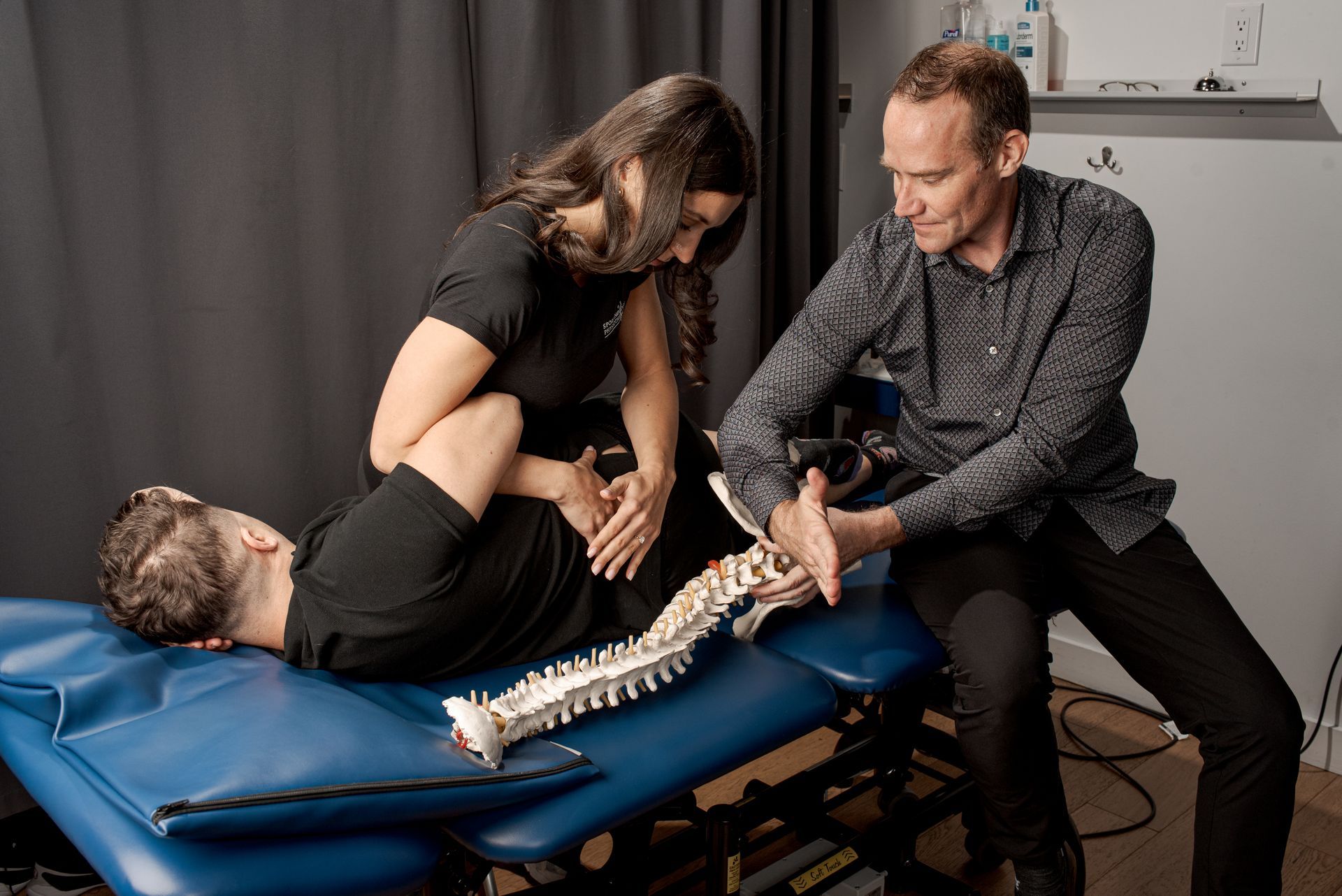A chiropractor instructs a professional while practicing a spinal adjustment on a patient lying on a clinic table.