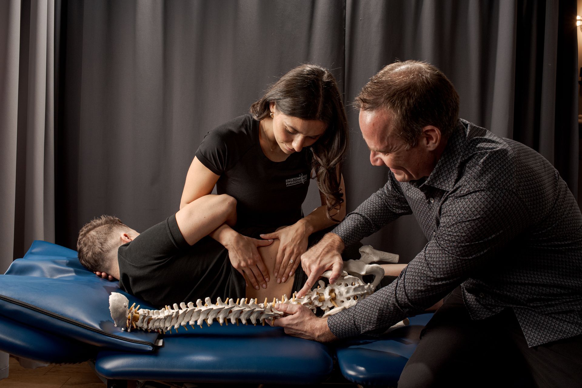 A professional showing a model of a human spine to a practitioner who is examining a patient lying on a treatment table.
