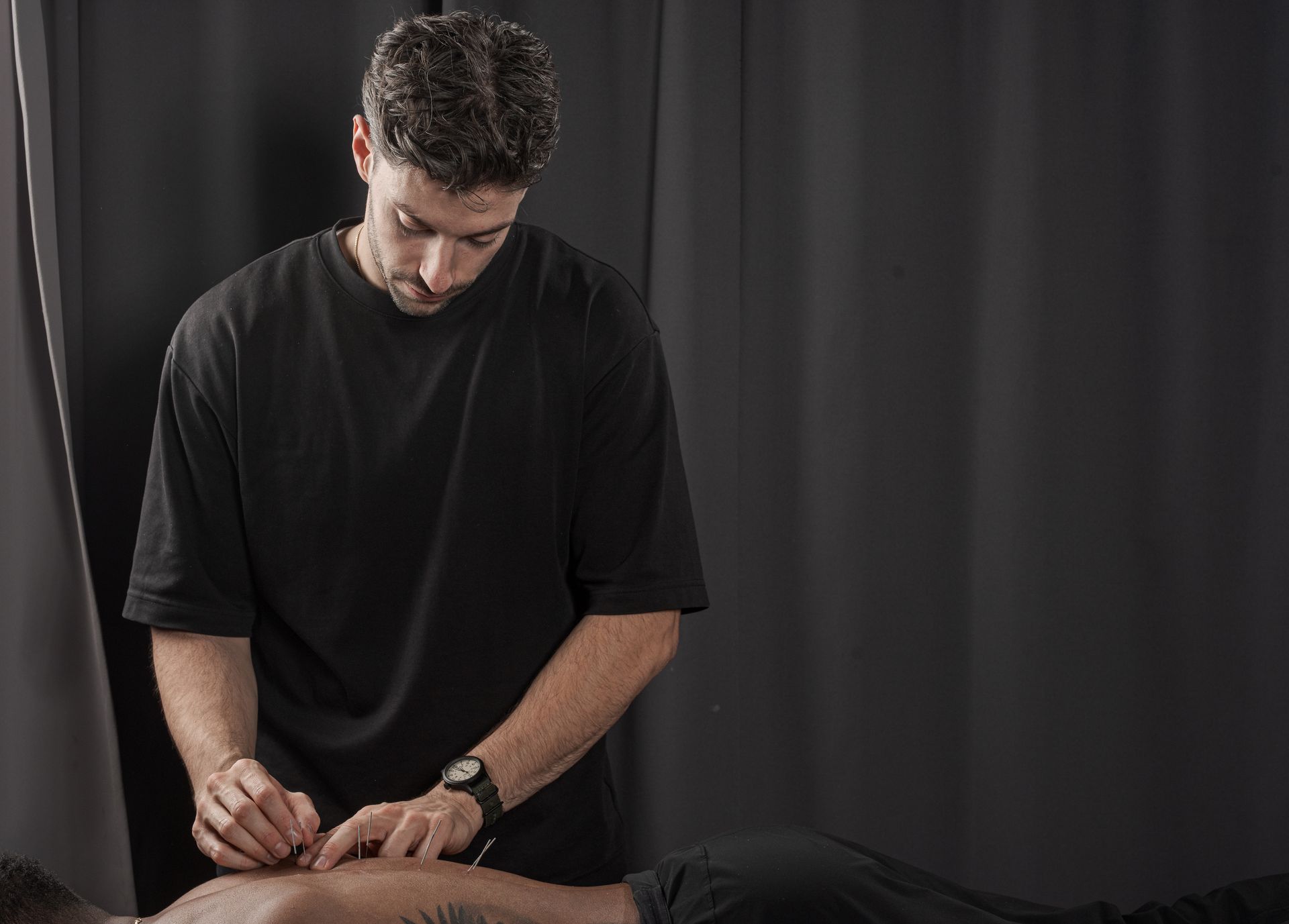 A practitioner in a black shirt performs dry needling therapy on a patient's back against a plain dark background.