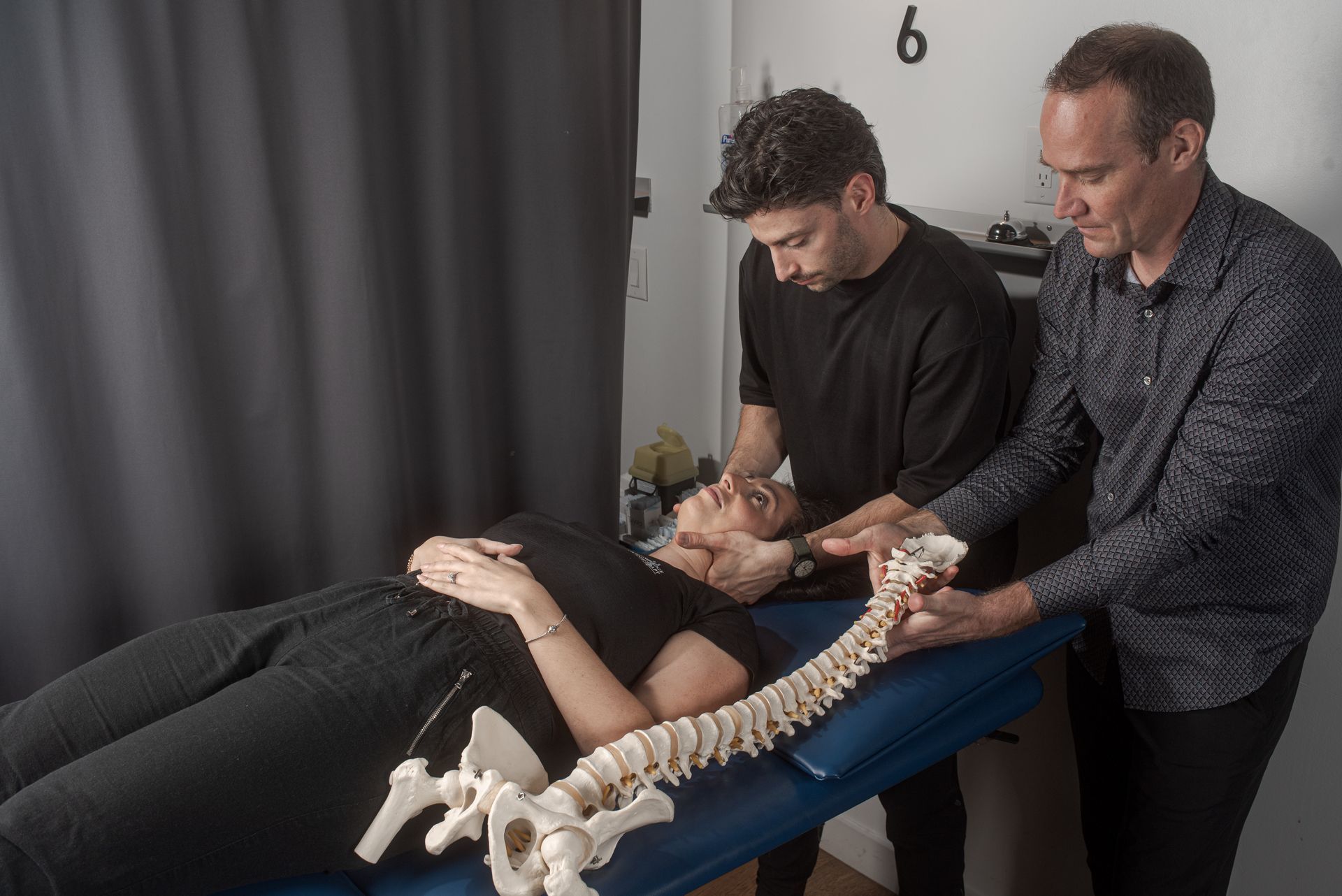 Two people demonstrate a chiropractic adjustment on a patient lying on a table next to a model of a human spine.