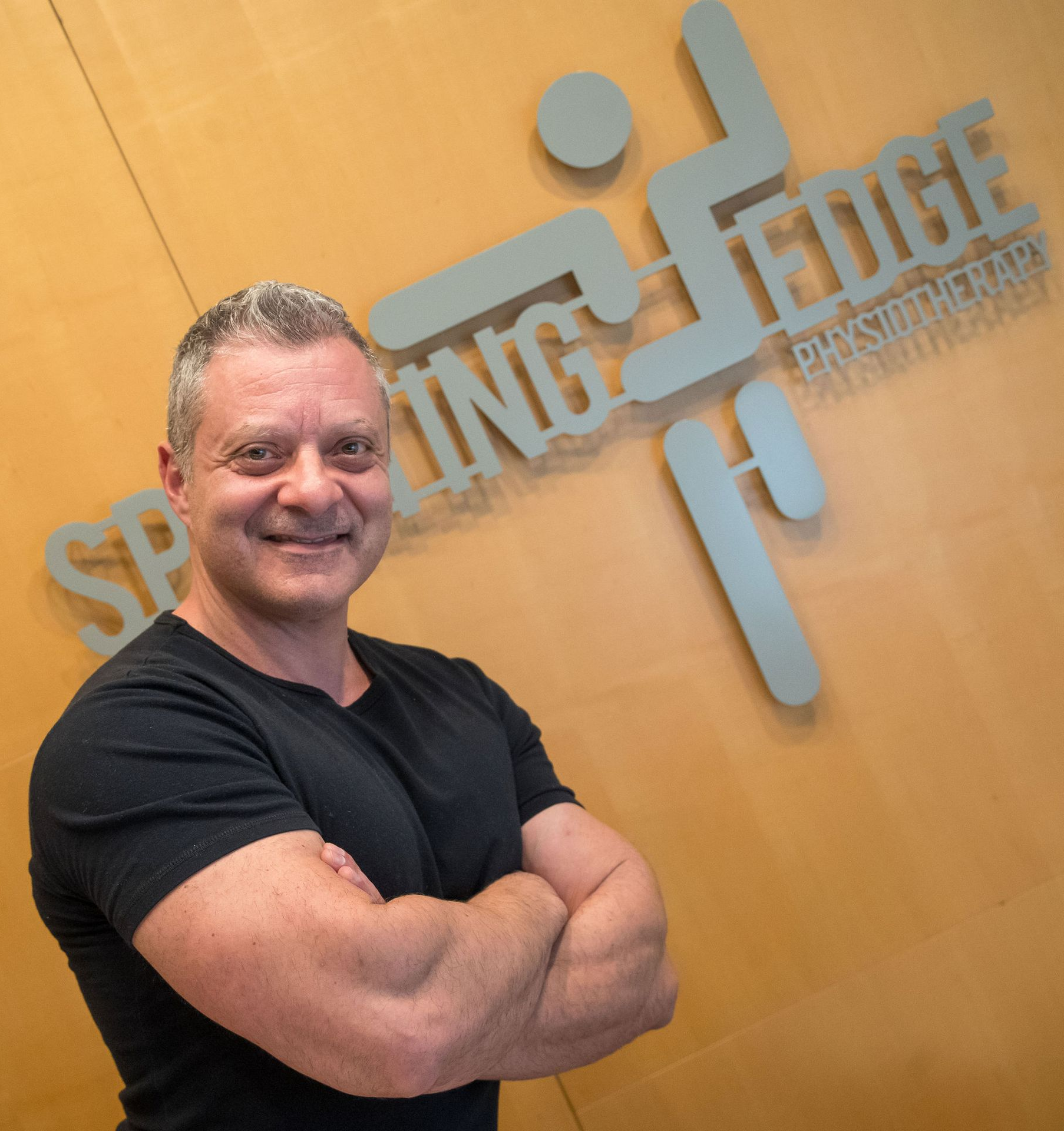 Manual Ostepathic Practitioner Anthony D'Assisi posing with arms crossed in front of a silver sporting edge physiotherapy sign