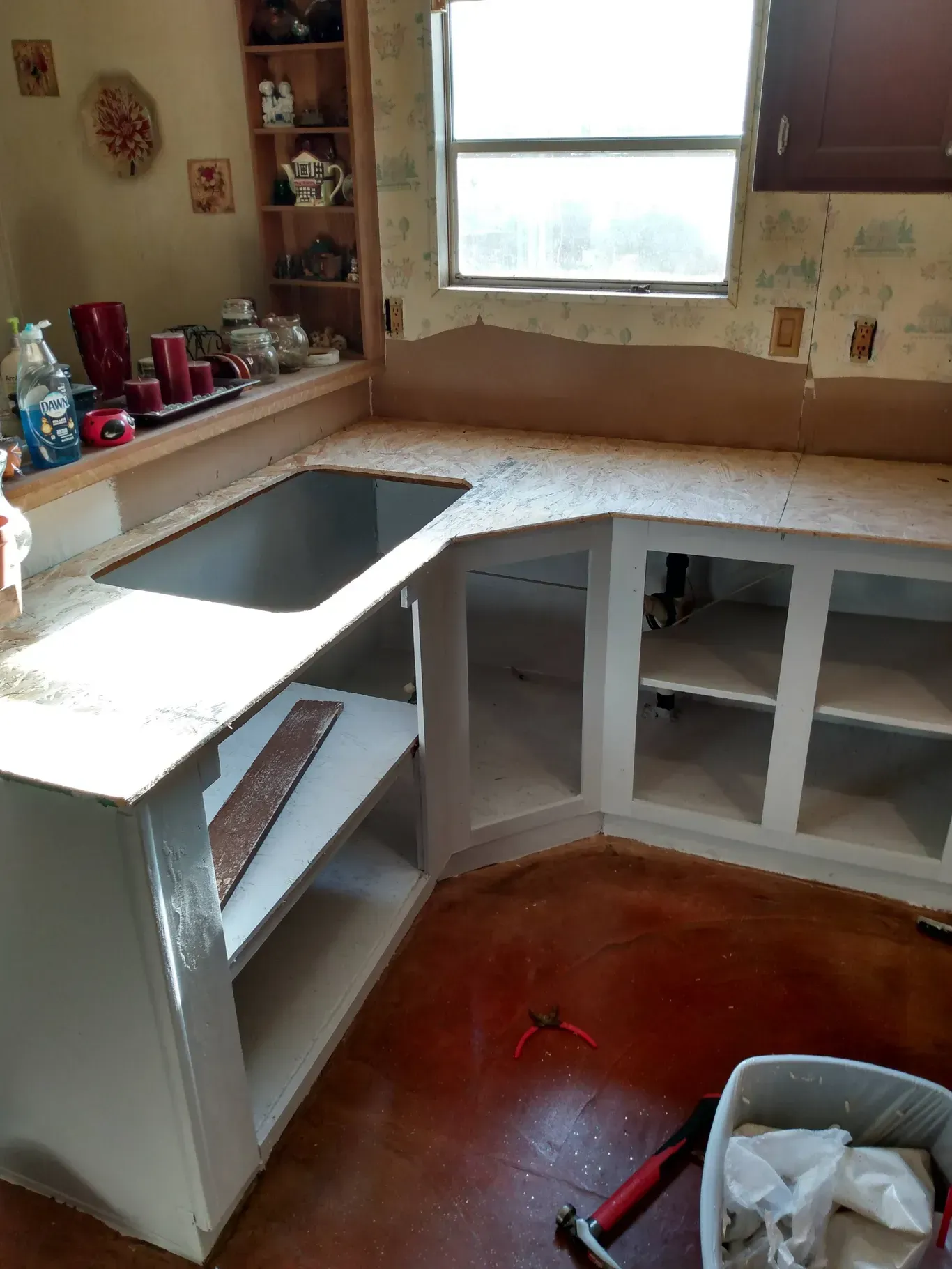 Kitchen under construction: white cabinets, wooden countertops, window, red floor, unfinished.