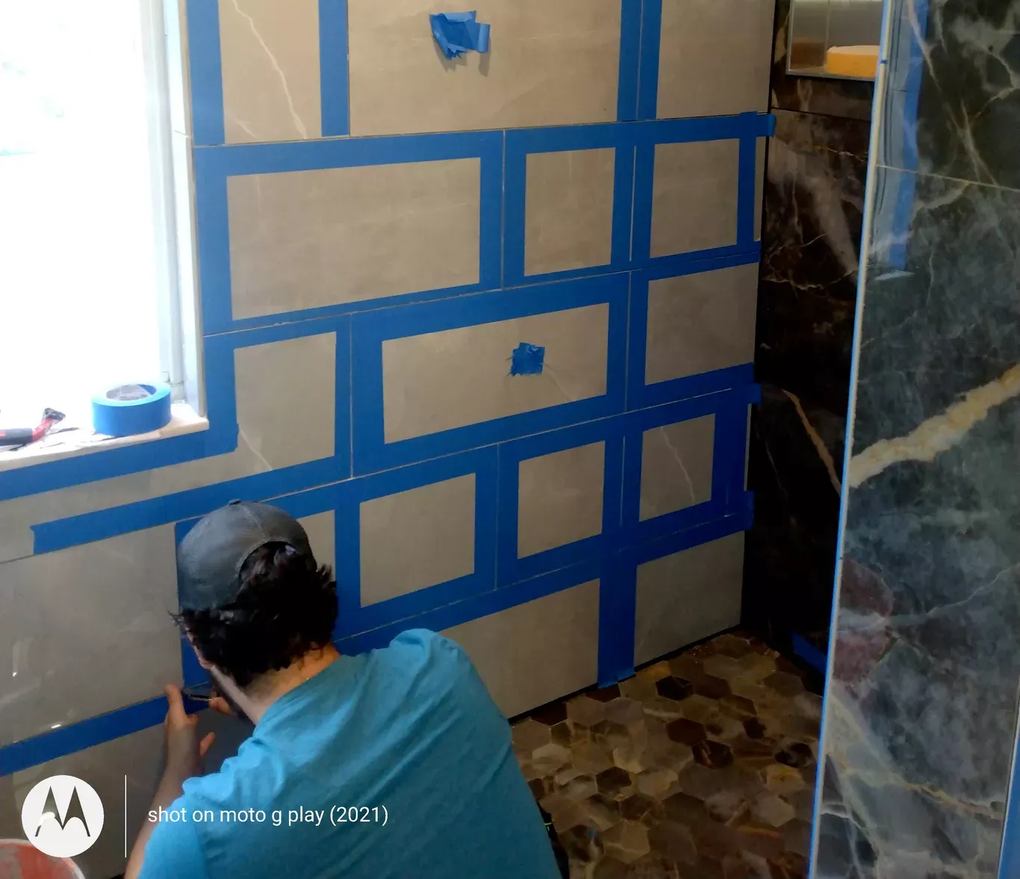 Man taping tiles in a bathroom. Blue tape outlines tile sections on gray wall. Window is on the left.