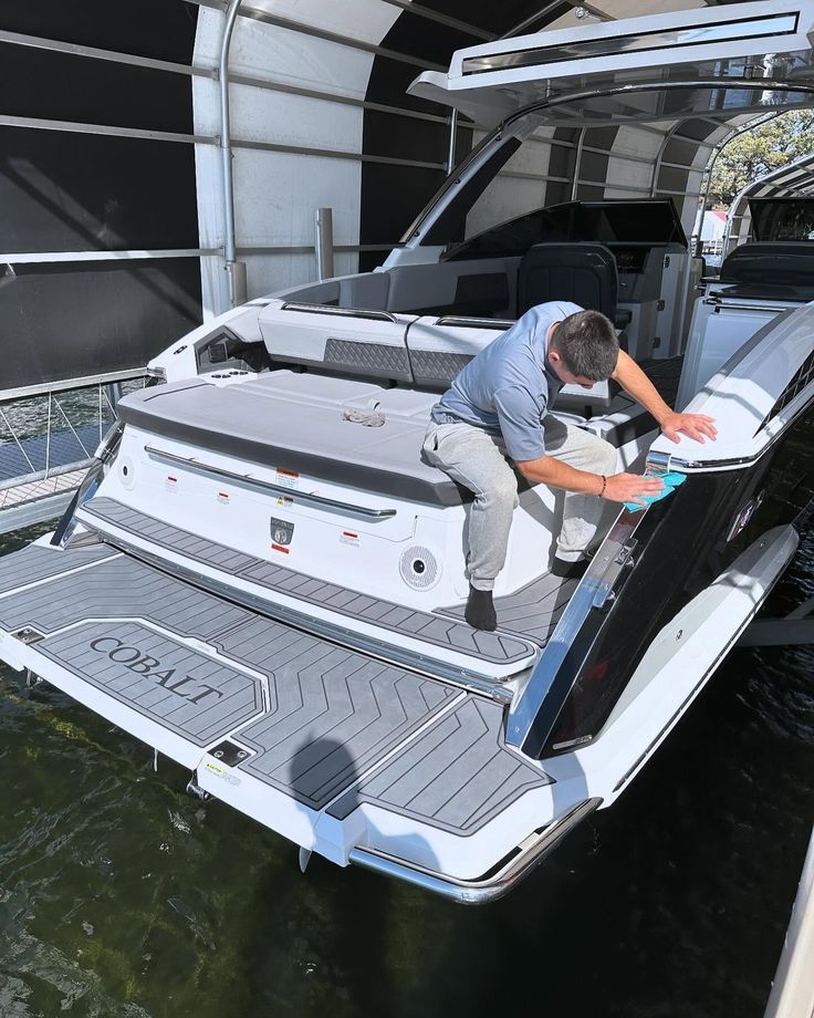 A man cleans a black and white motorboat's rear, docked near a canopy. 