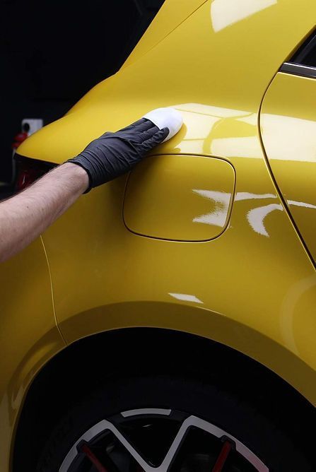 Person in black gloves applying wax to a yellow car's body panel with a white applicator.