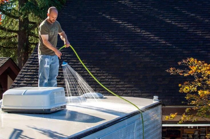 Man washing the roof of an RV with a hose outdoors, near a house with a dark roof and trees.