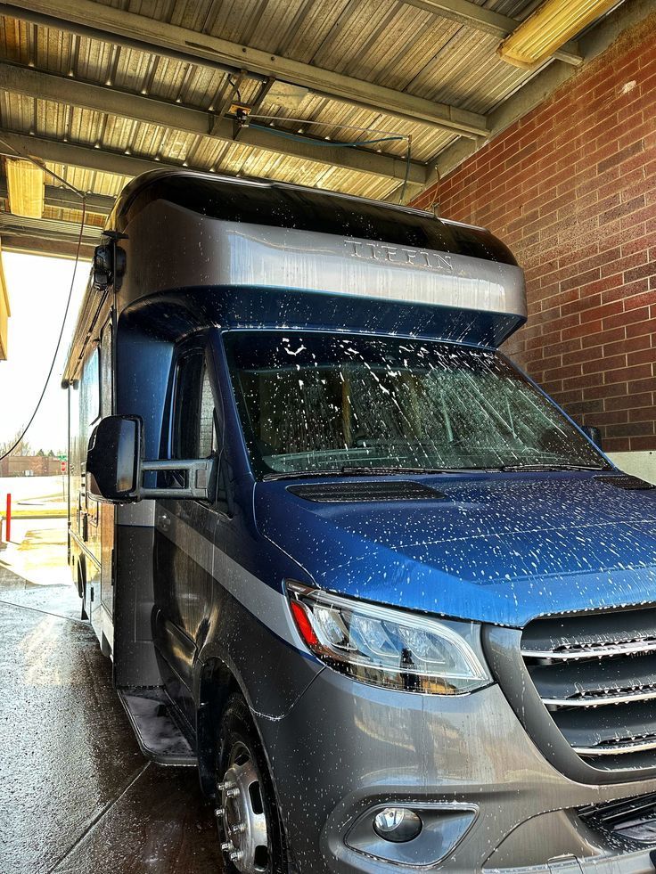 Blue semi-truck cab being washed under a covered bay, with water spraying on the windshield.