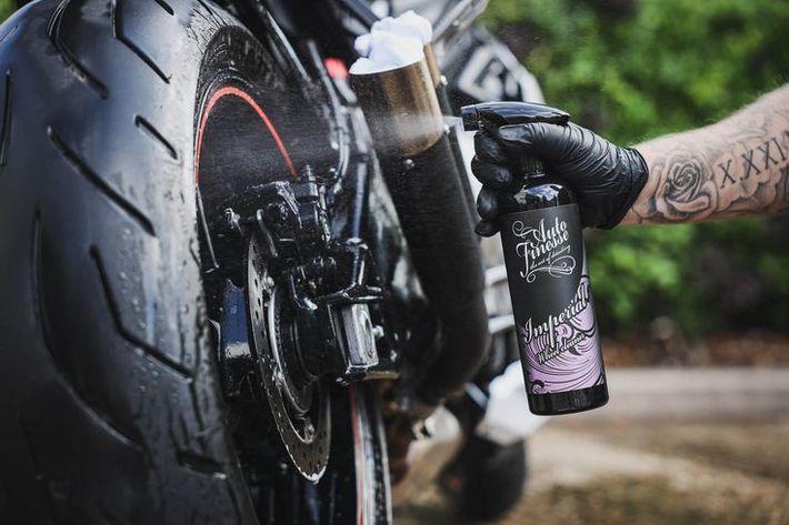 Hand gloved on motorcycle chain, holding a black and purple cleaning spray bottle beside the rear wheel.