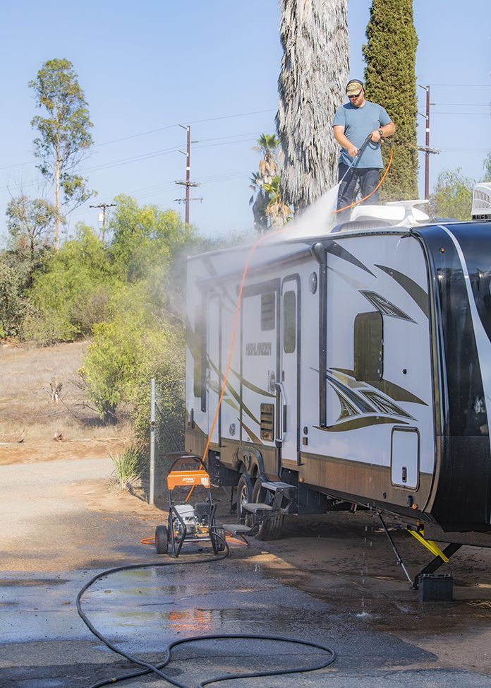 Man power washes the roof of a camper trailer. Orange pressure washer, green trees, sunny outdoors.