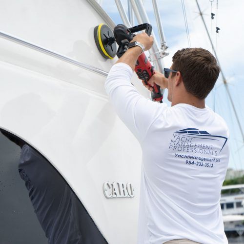 Man polishing a white boat hull with a power buffer near the water.