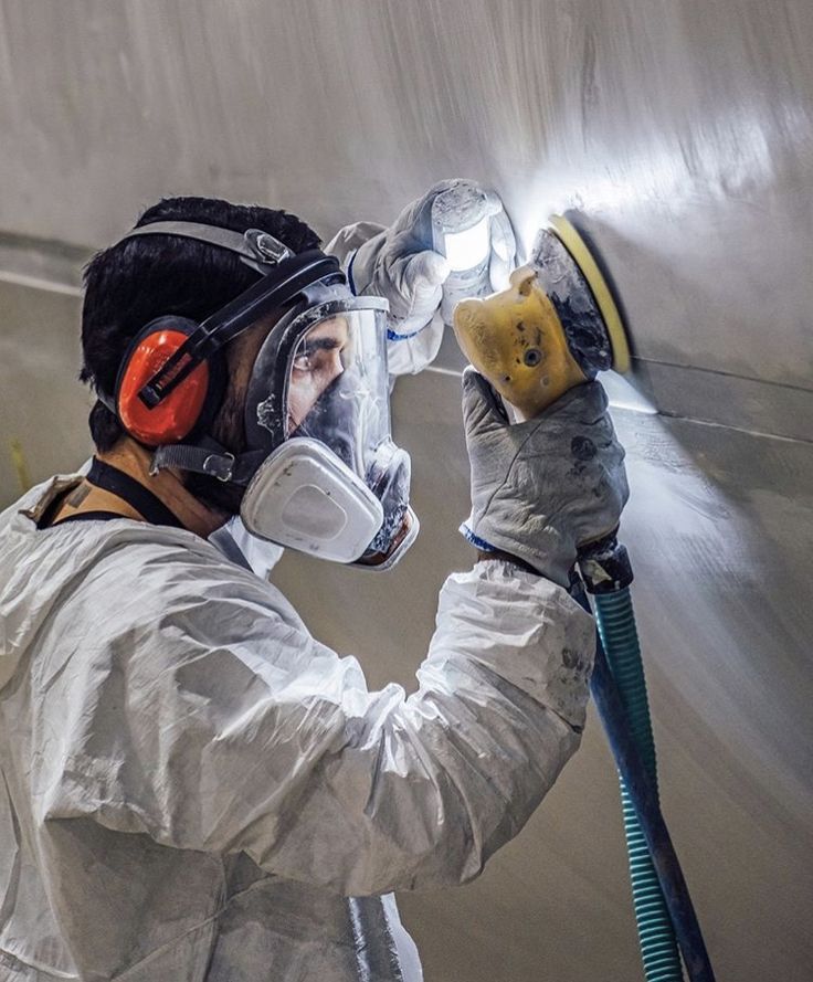Person in protective gear sanding a surface, illuminated by a light.