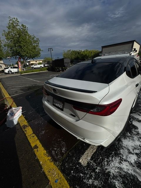 White sedan in a wet parking lot, rear view with foam on the ground under cloudy skies